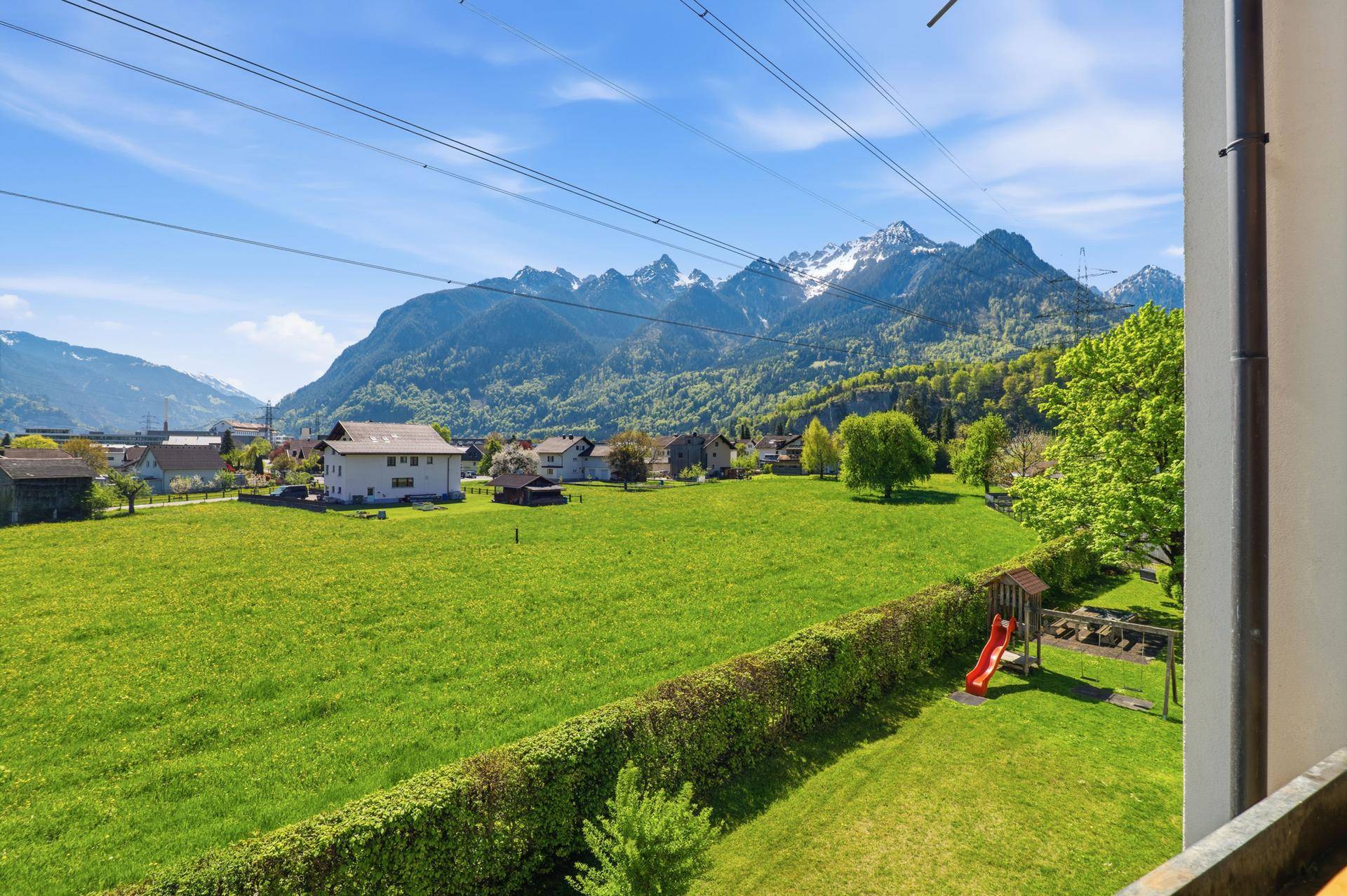 Grüne Wiese mit Kinderspielplatz, Hecke und beeindruckendem Bergpanorama im Hintergrund.