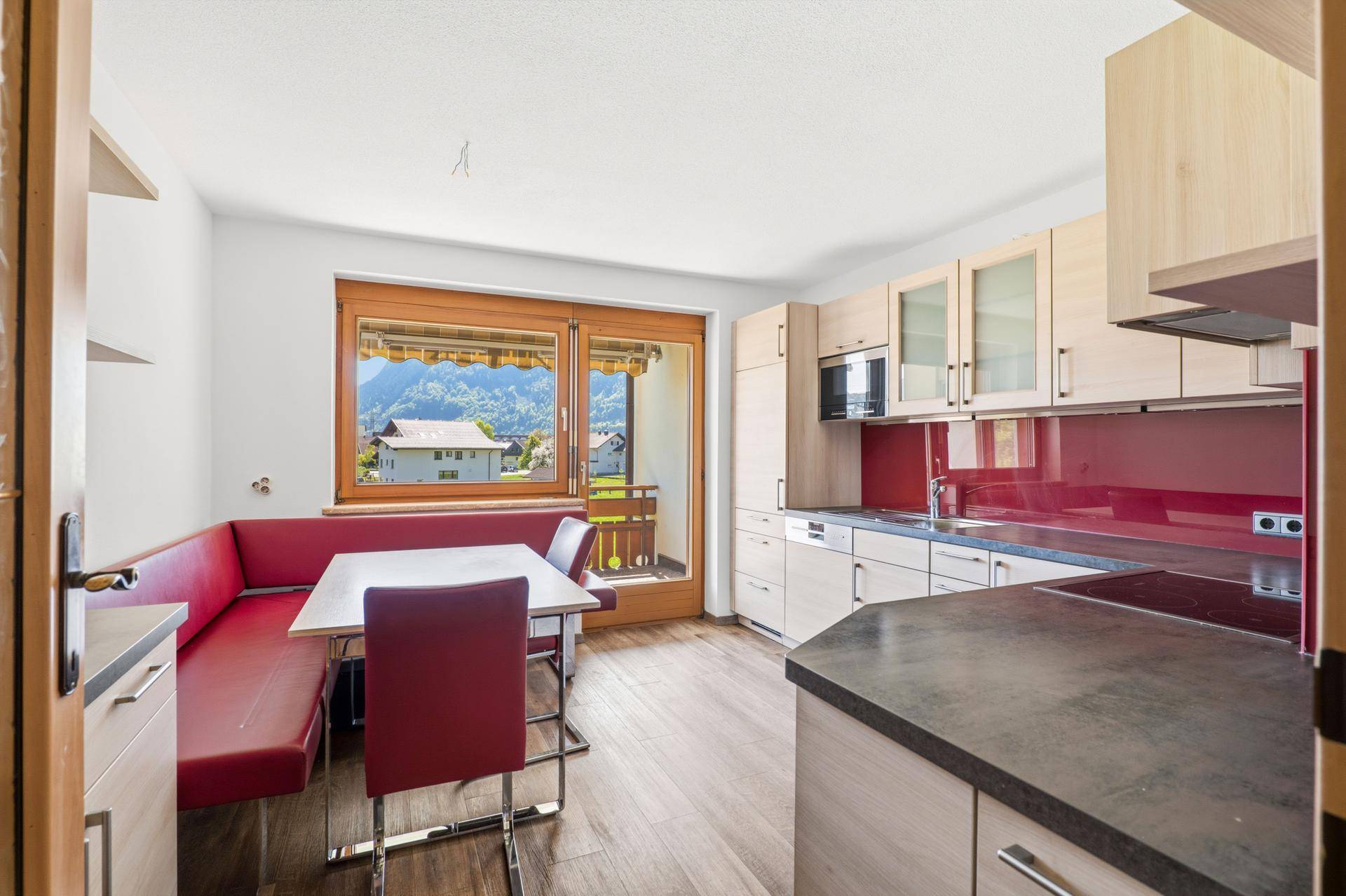 Einbauküche mit Essecke, roten Akzenten und Zugang zum Balkon mit Bergblick.