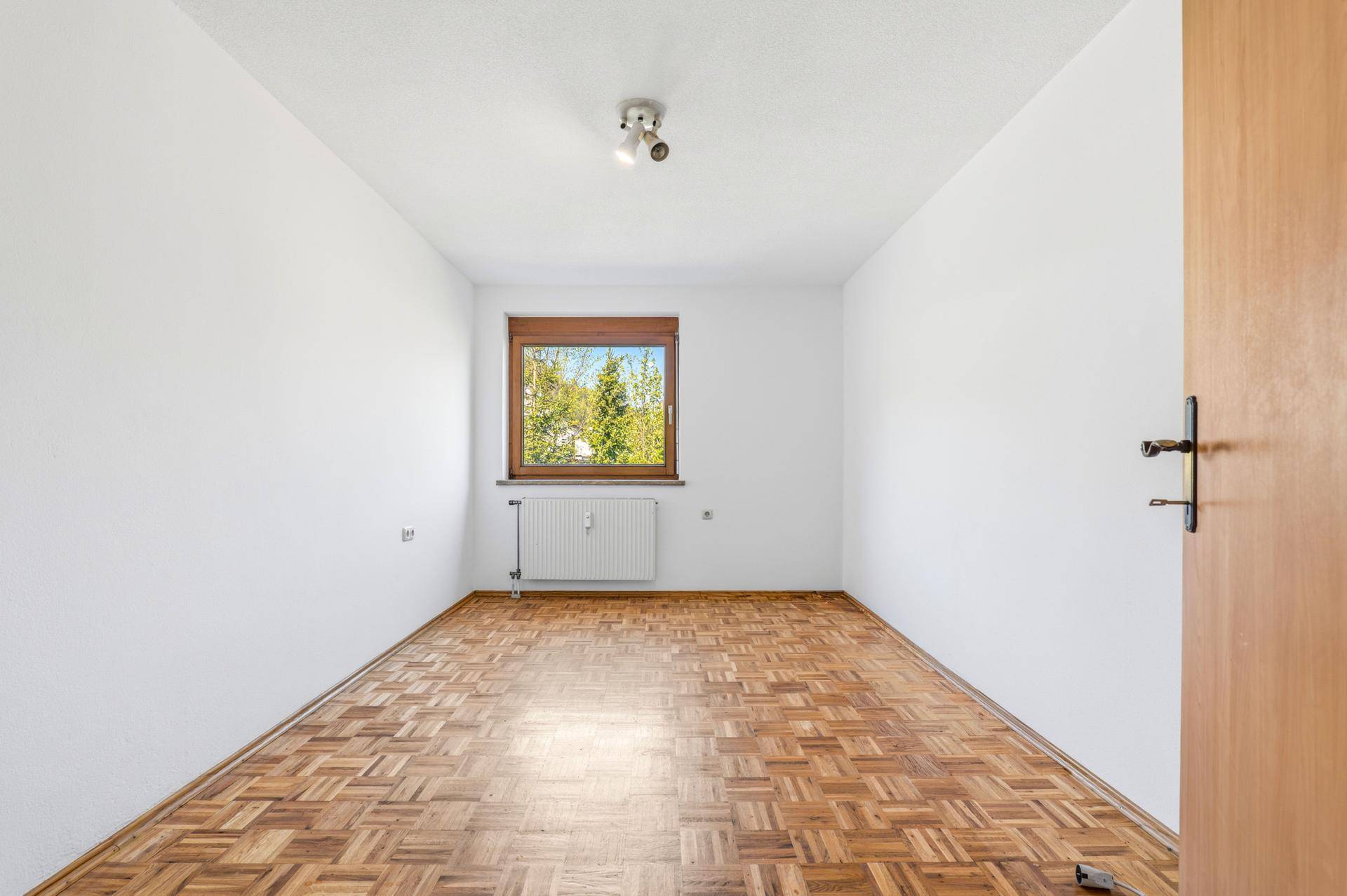 Helles Schlafzimmer mit Parkettboden, Heizkörper unter dem Fenster und Blick ins Grüne.