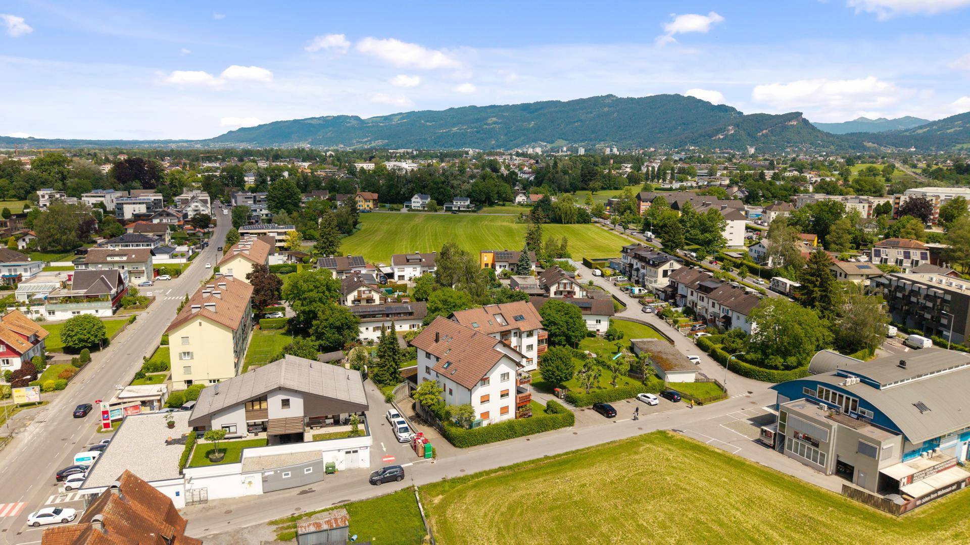 Luftaufnahme einer Wohngegend mit Häusern, Feldern und Bergen im Hintergrund.