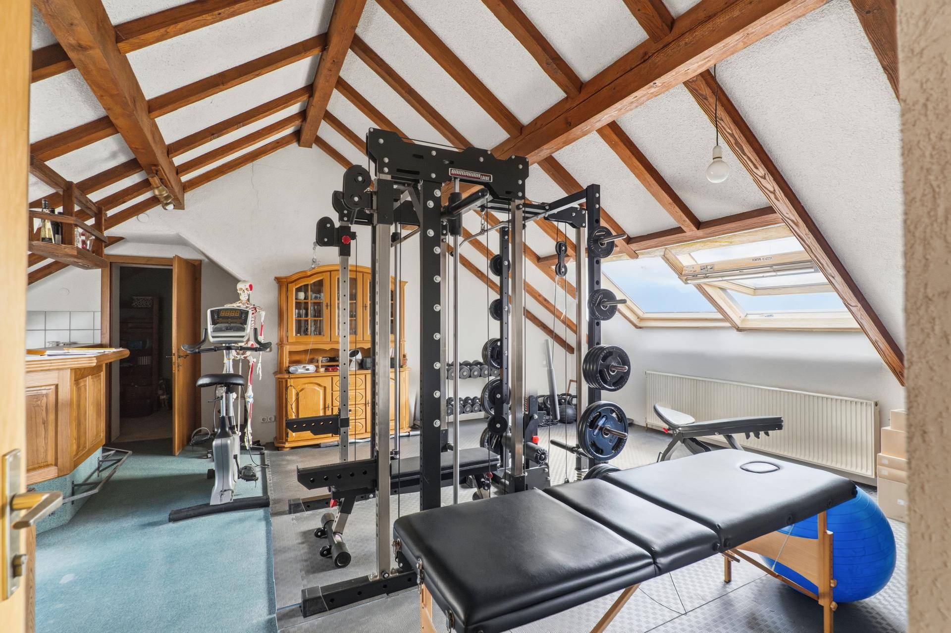 Fitnessraum mit verschiedenen Trainingsgeräten, Holzbalken und Dachfenstern.