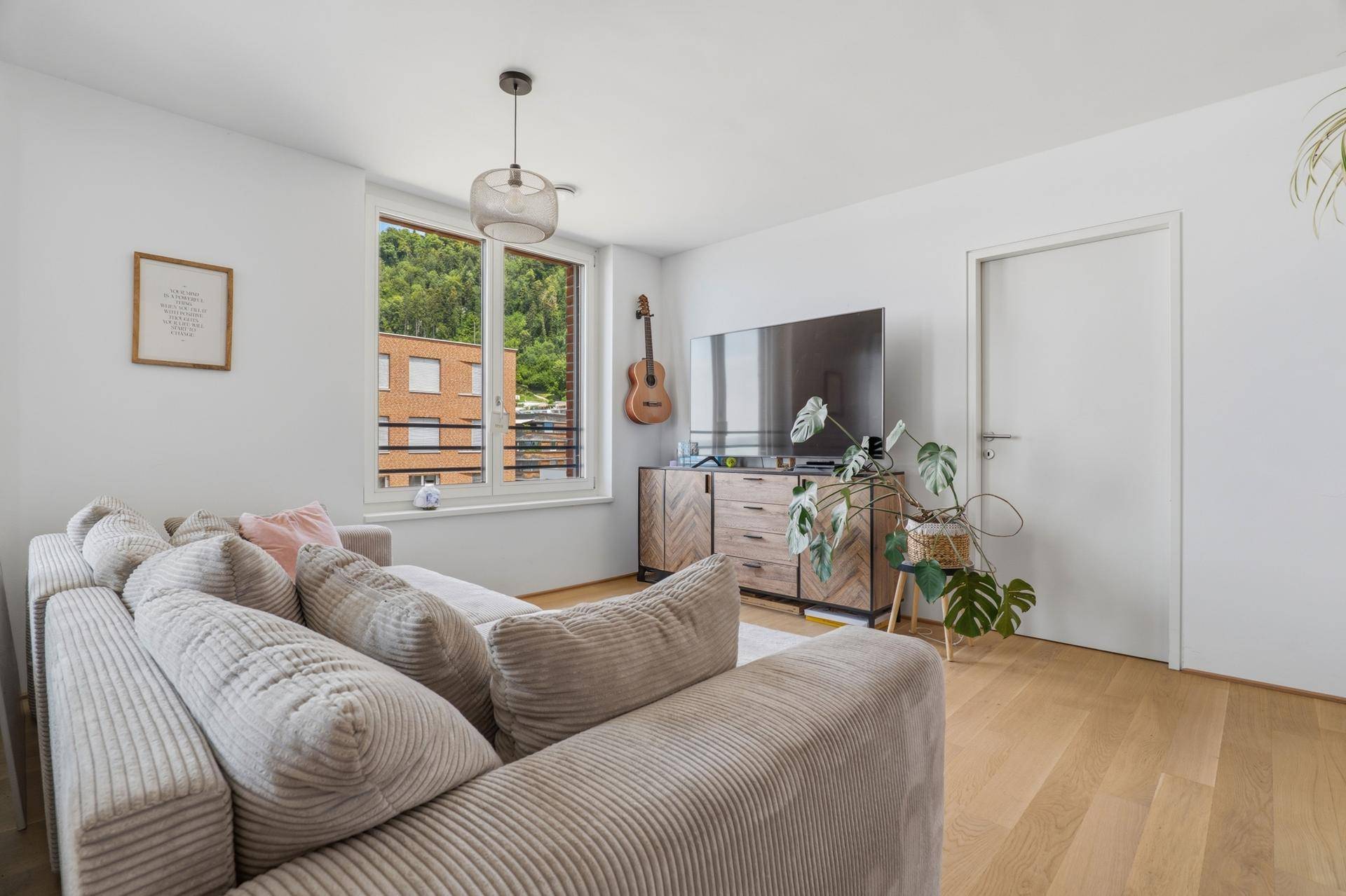 Gemütliches Wohnzimmer mit Sofa, Fernseher und Blick ins Grüne durch große Fenster.