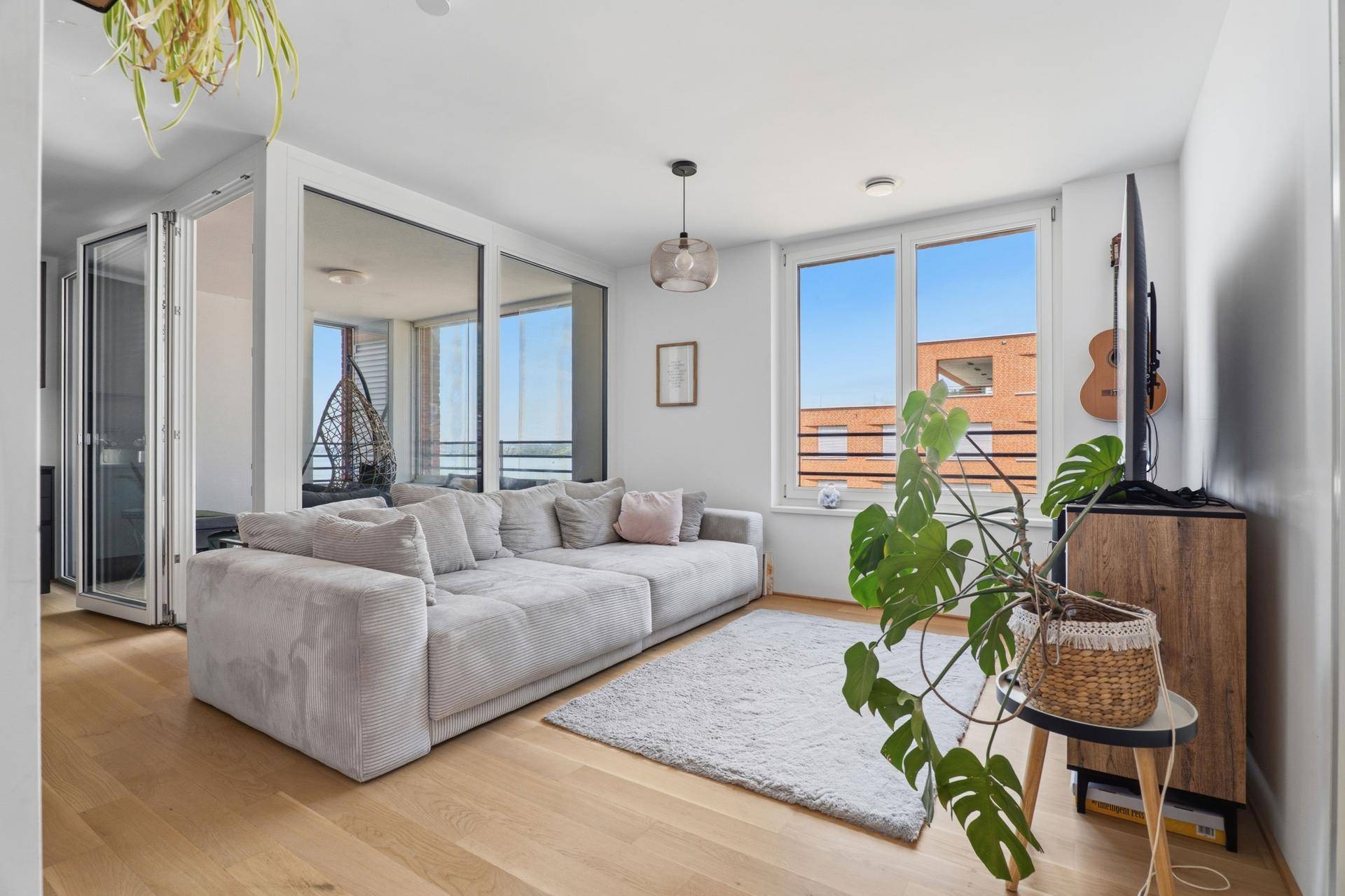 Helles Wohnzimmer mit großem Sofa, Holzboden und Zugang zu einem verglasten Balkon.