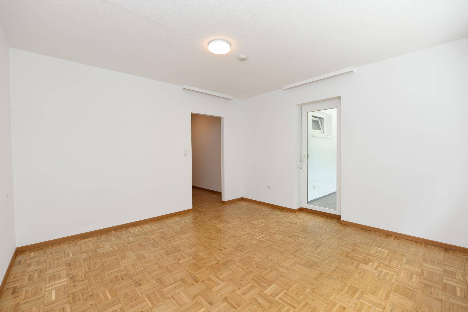 Heller, leerer Raum mit Parkettboden und Zugang zu einem weiteren Zimmer sowie zum Balkon.