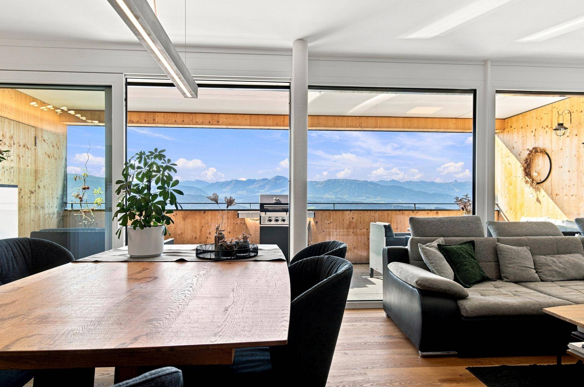 Heller Wohn- und Essbereich mit Holztisch, bequemer Couch und Panoramablick auf die Alpen durch die Fensterfront.