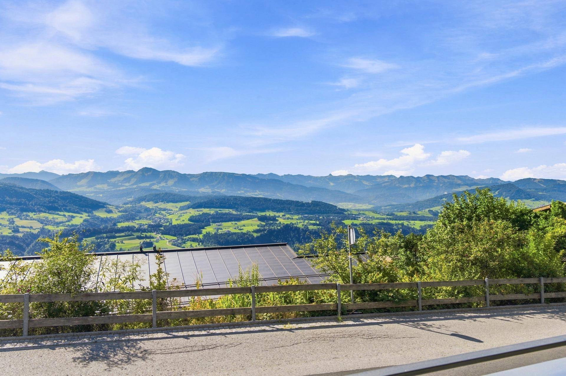 Weitläufiger Blick auf die grüne Alpenlandschaft mit bewaldeten Hügeln und einem Gebäude mit Solaranlage.