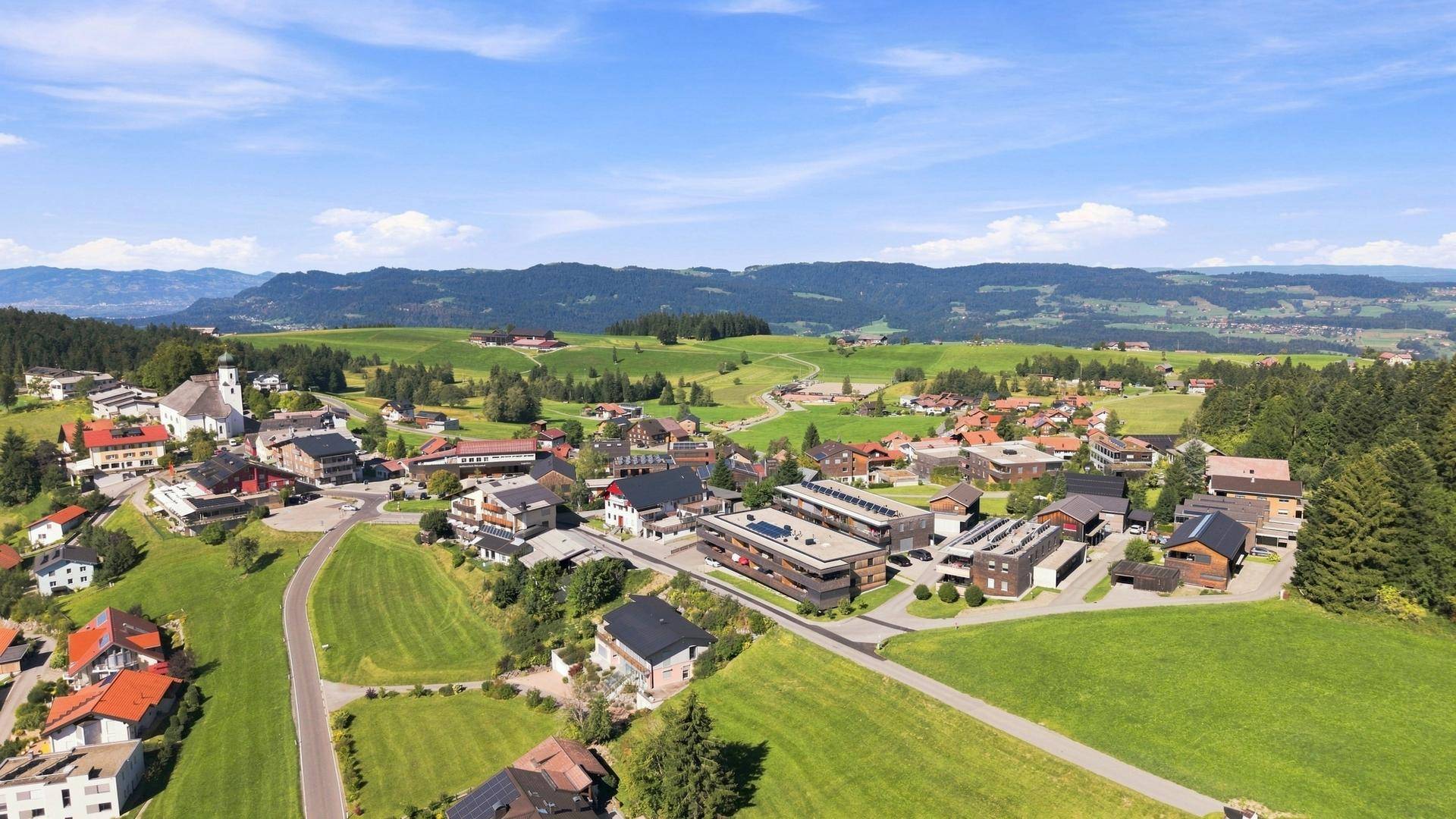 Luftaufnahme eines charmanten Dorfes mit traditionellen und modernen Gebäuden, umgeben von grünen Wiesen.