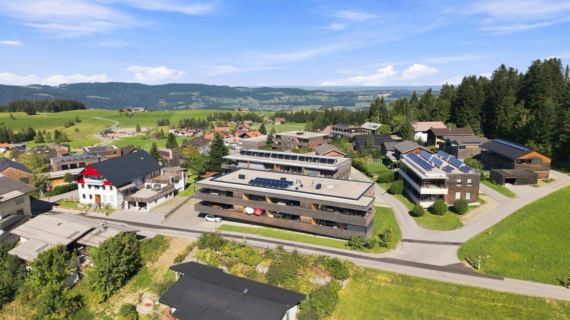 Weitläufige Luftaufnahme eines modernen Wohnkomplexes in einer idyllischen grünen Landschaft mit Bergen im Hintergrund.