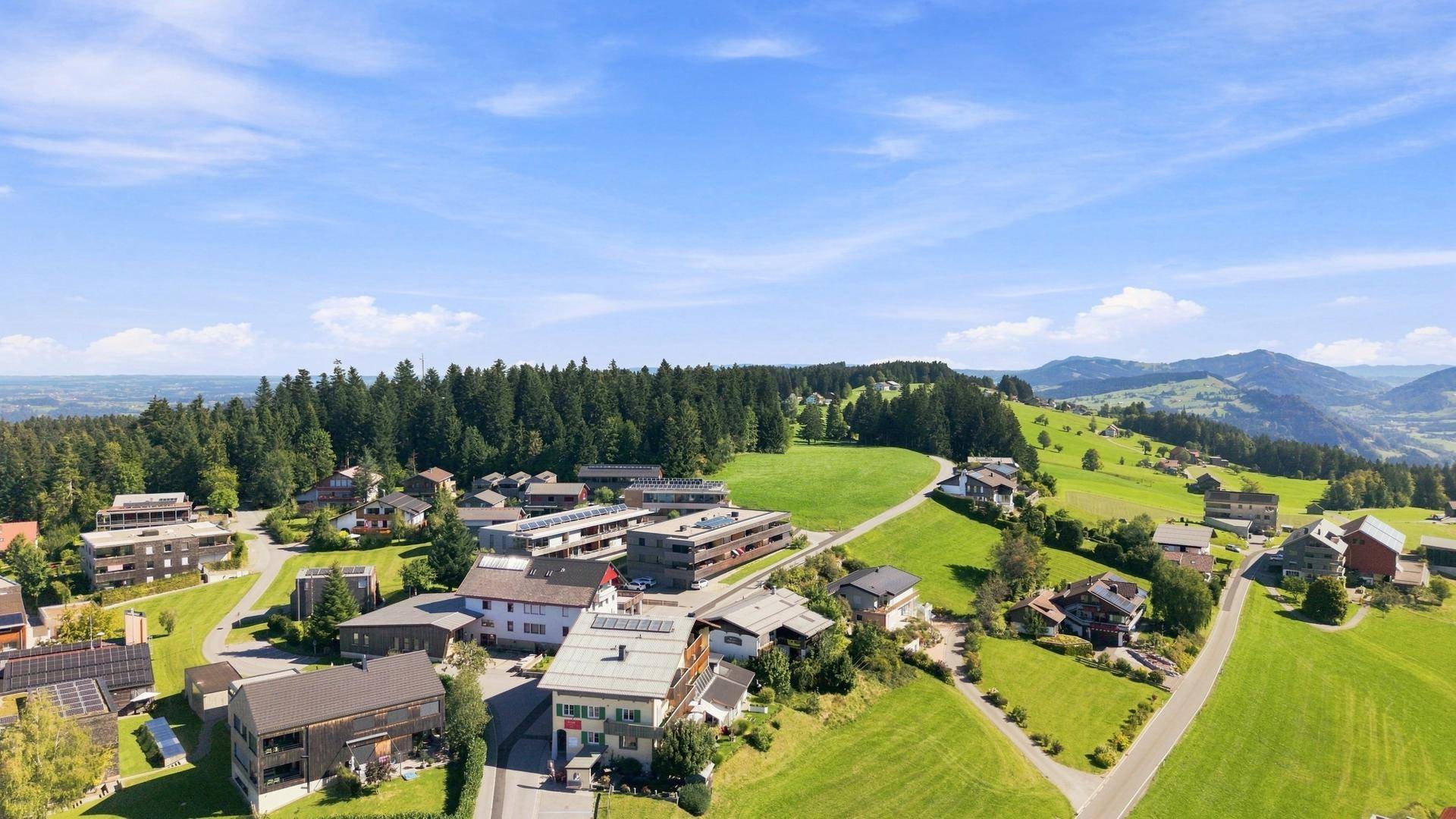 Weitläufige Luftaufnahme einer ländlichen Siedlung, eingebettet in eine hügelige, bewaldete Landschaft unter blauem Himmel.