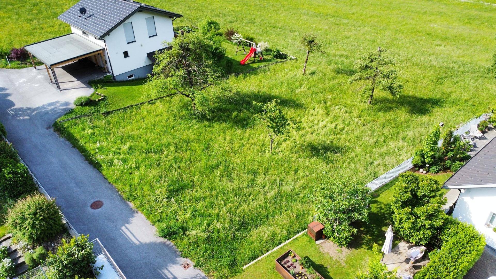 Weitläufiges grünes Grundstück mit angrenzender Straße und einem Haus mit Carport im Hintergrund.