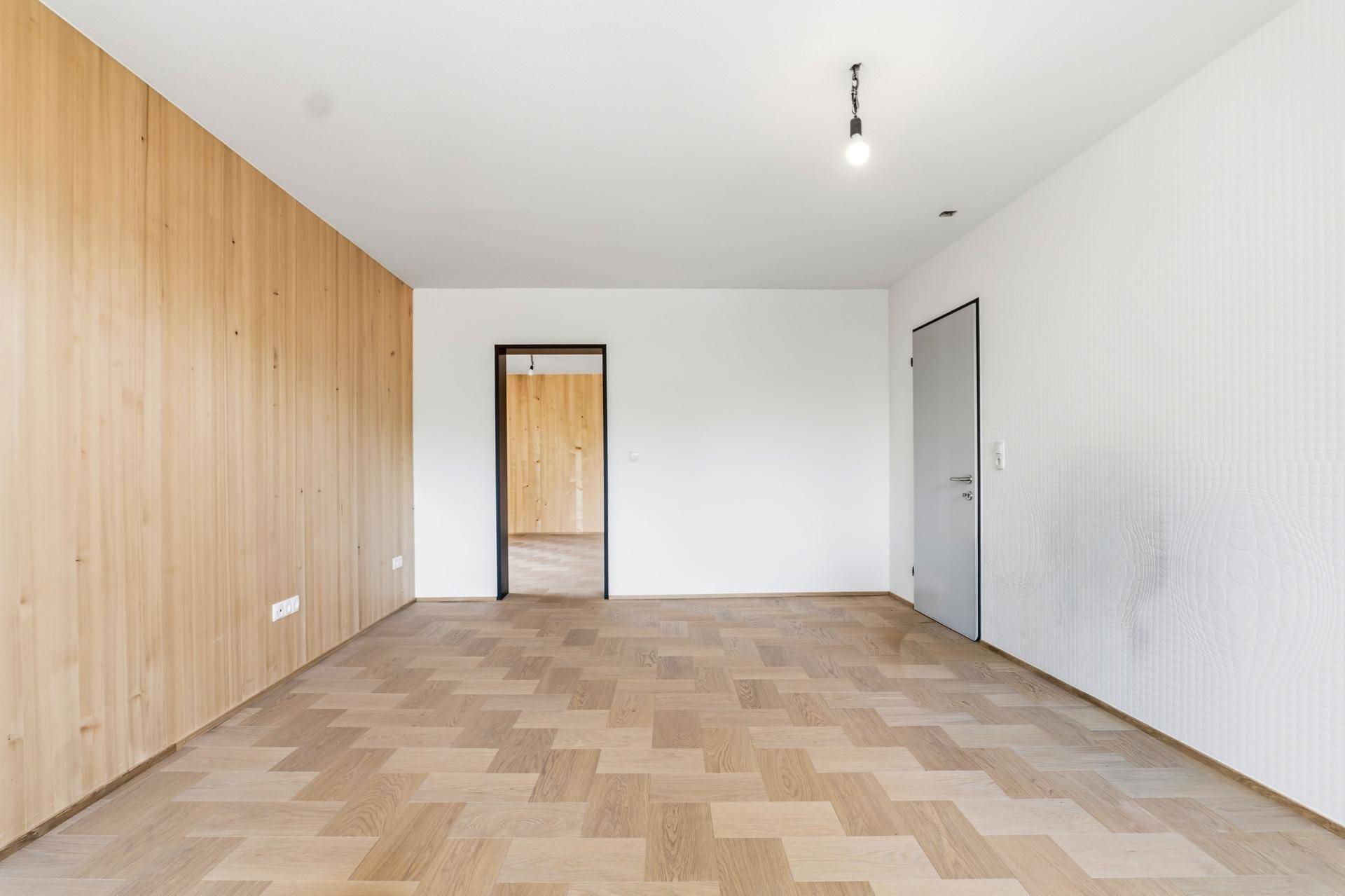 Leerer Raum mit Fischgrätparkett, Holzwandverkleidung und Zugang zu einem weiteren Zimmer.
