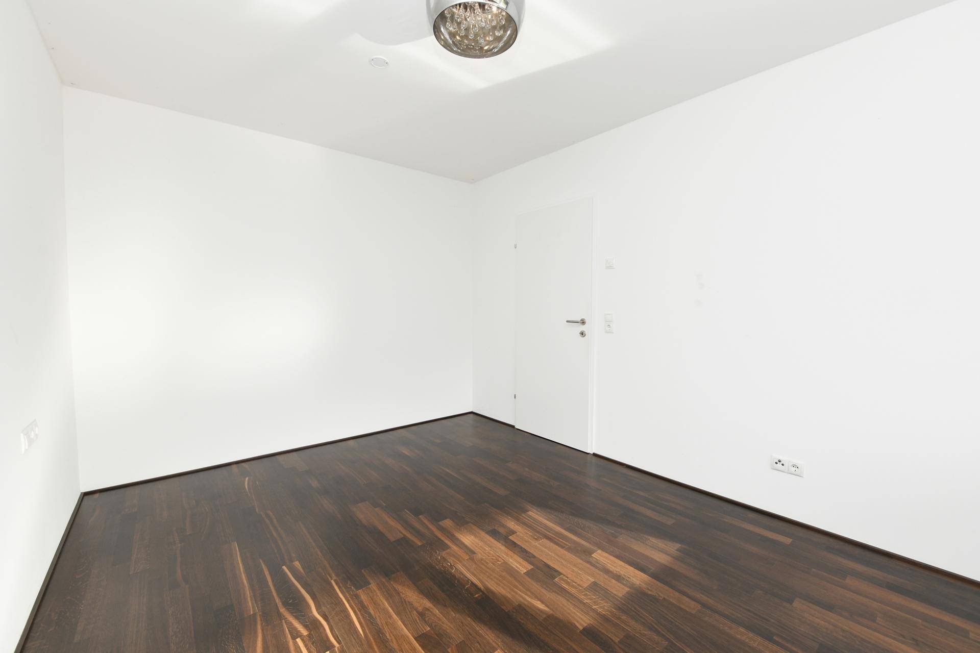 Leeres Schlafzimmer mit dunklem Holzboden und weißen Wänden.