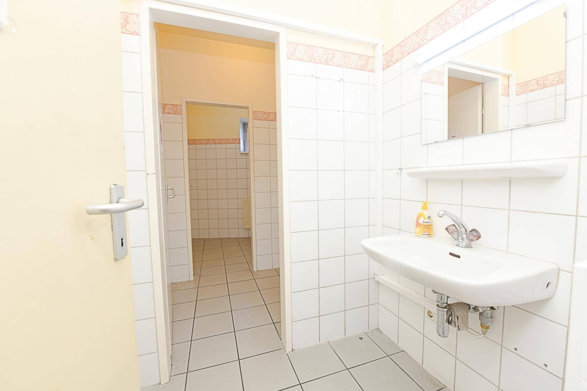Badezimmer mit weiß gefliesten Wänden, Waschbecken und Zugang zu einem weiteren Raum, funktional.
