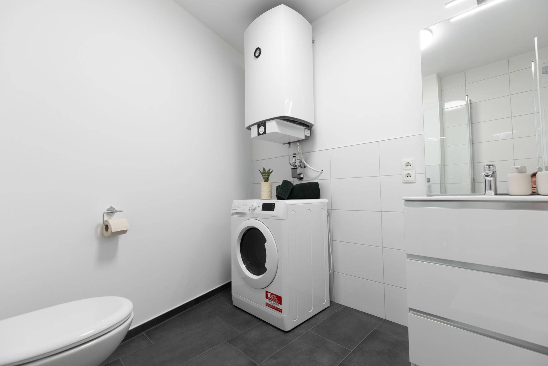 Funktionales Badezimmer mit WC, Waschmaschine, Warmwasserboiler und Waschtisch, praktisch eingerichtet.