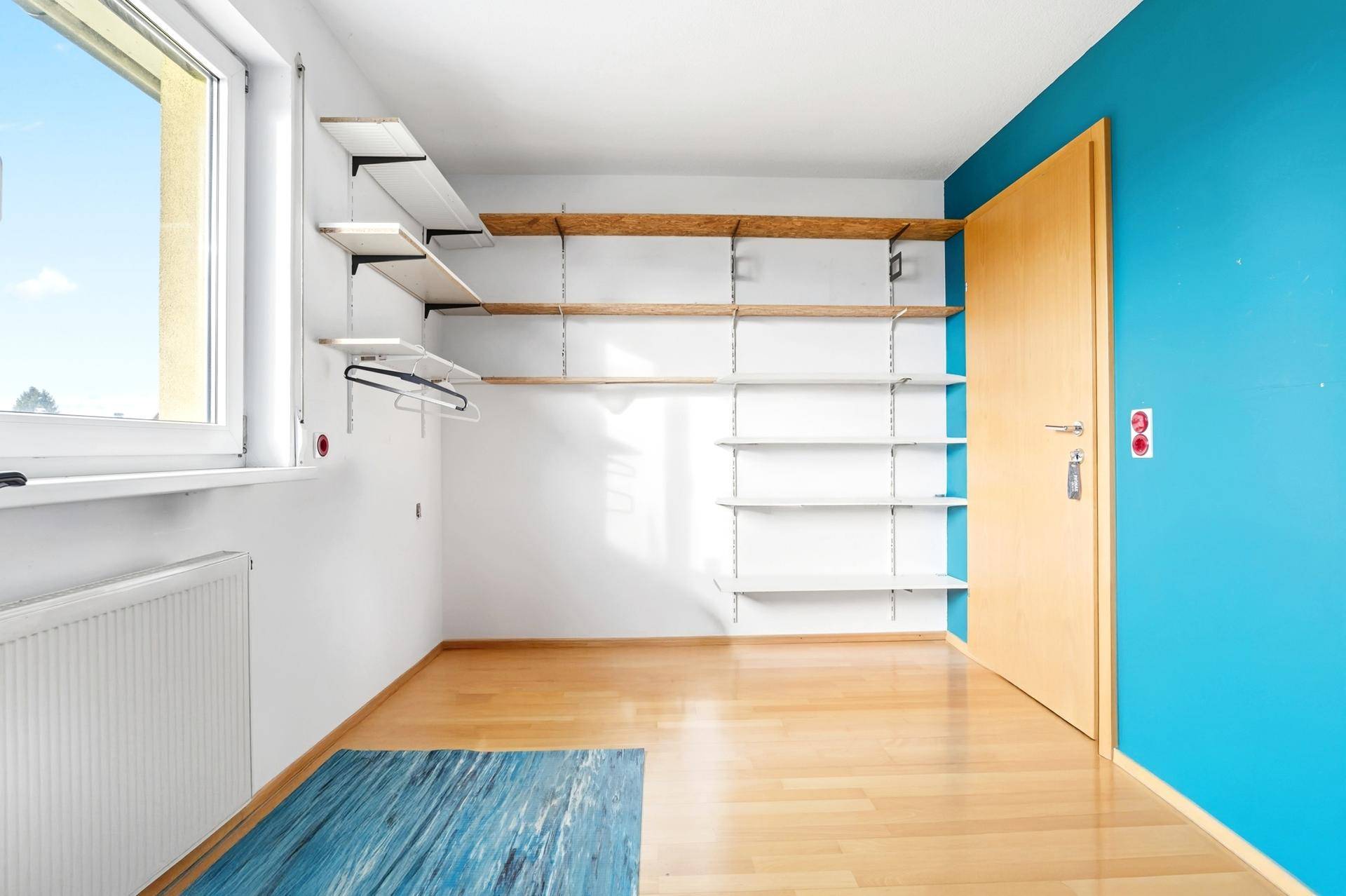 Helles Zimmer mit Holzboden, Regalsystem und Fenster, ideal als Ankleidezimmer.