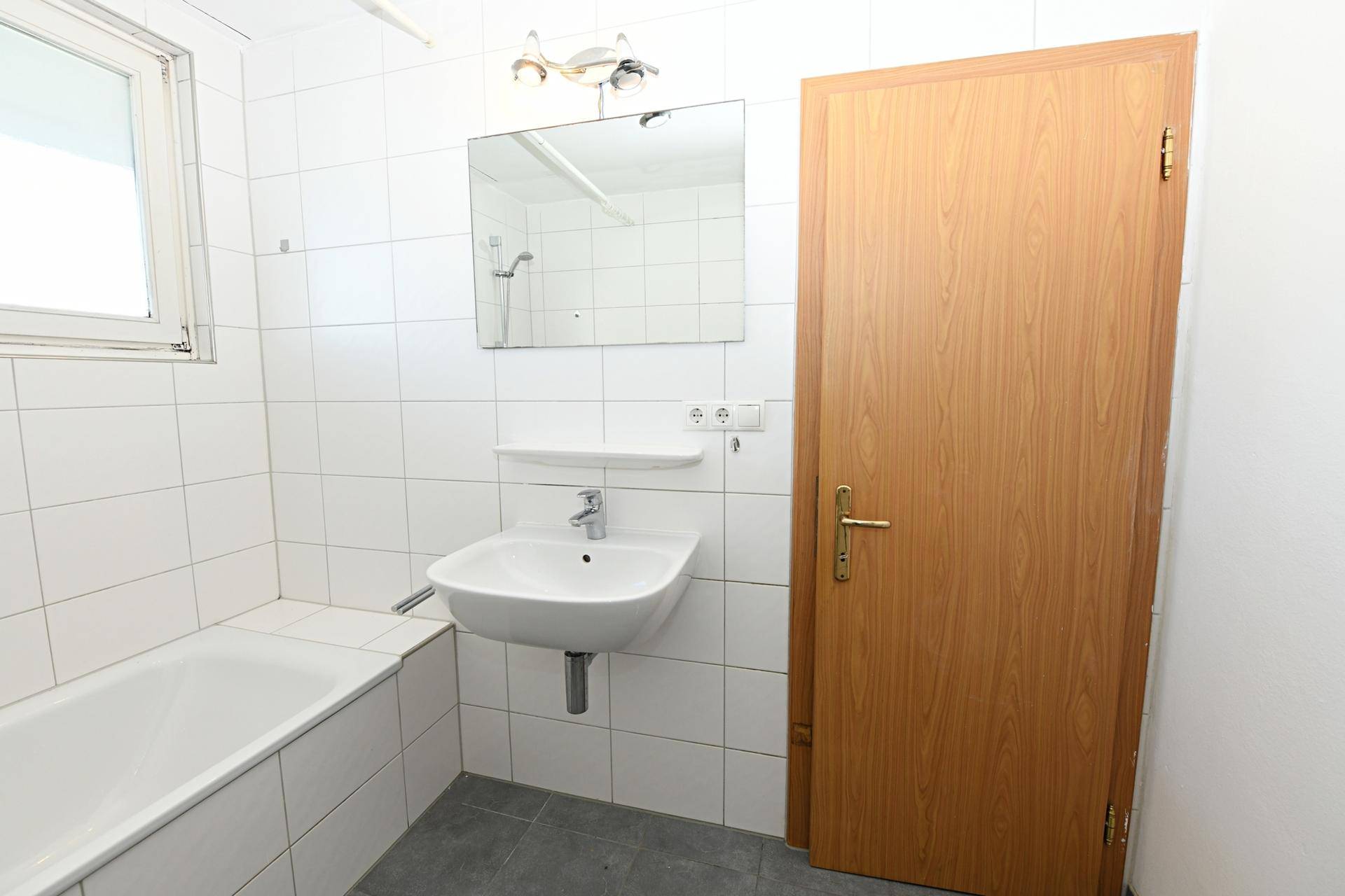 Helles Badezimmer mit weißen Wandfliesen, Waschbecken, Spiegel und Holztür.