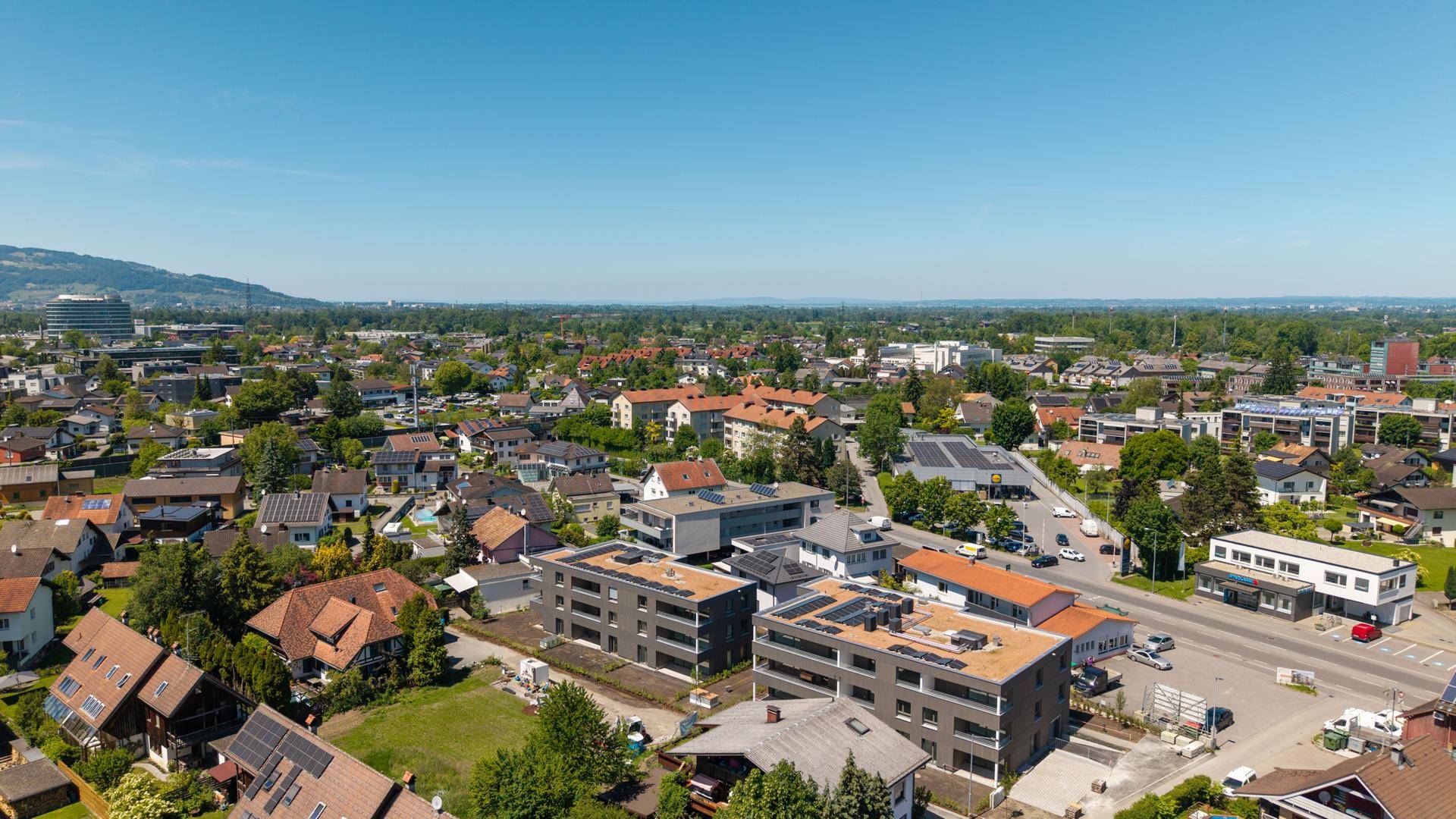 Luftaufnahme einer städtischen Wohngegend mit modernen Gebäuden und grüner Umgebung vor einer Bergkulisse.