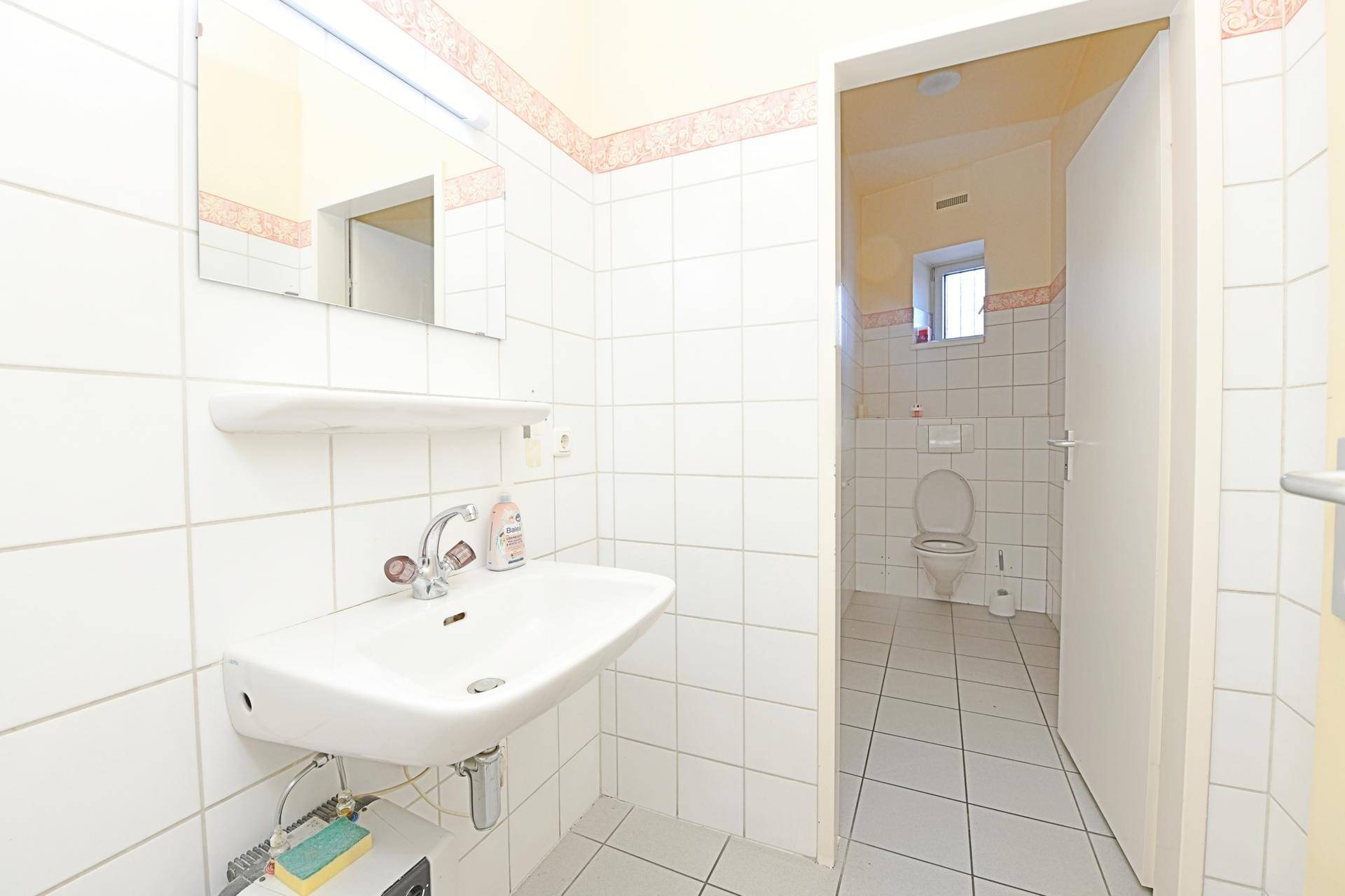 Badezimmer mit weiß gefliesten Wänden, Waschbecken und Spiegel, in gutem und sauberem Zustand.
