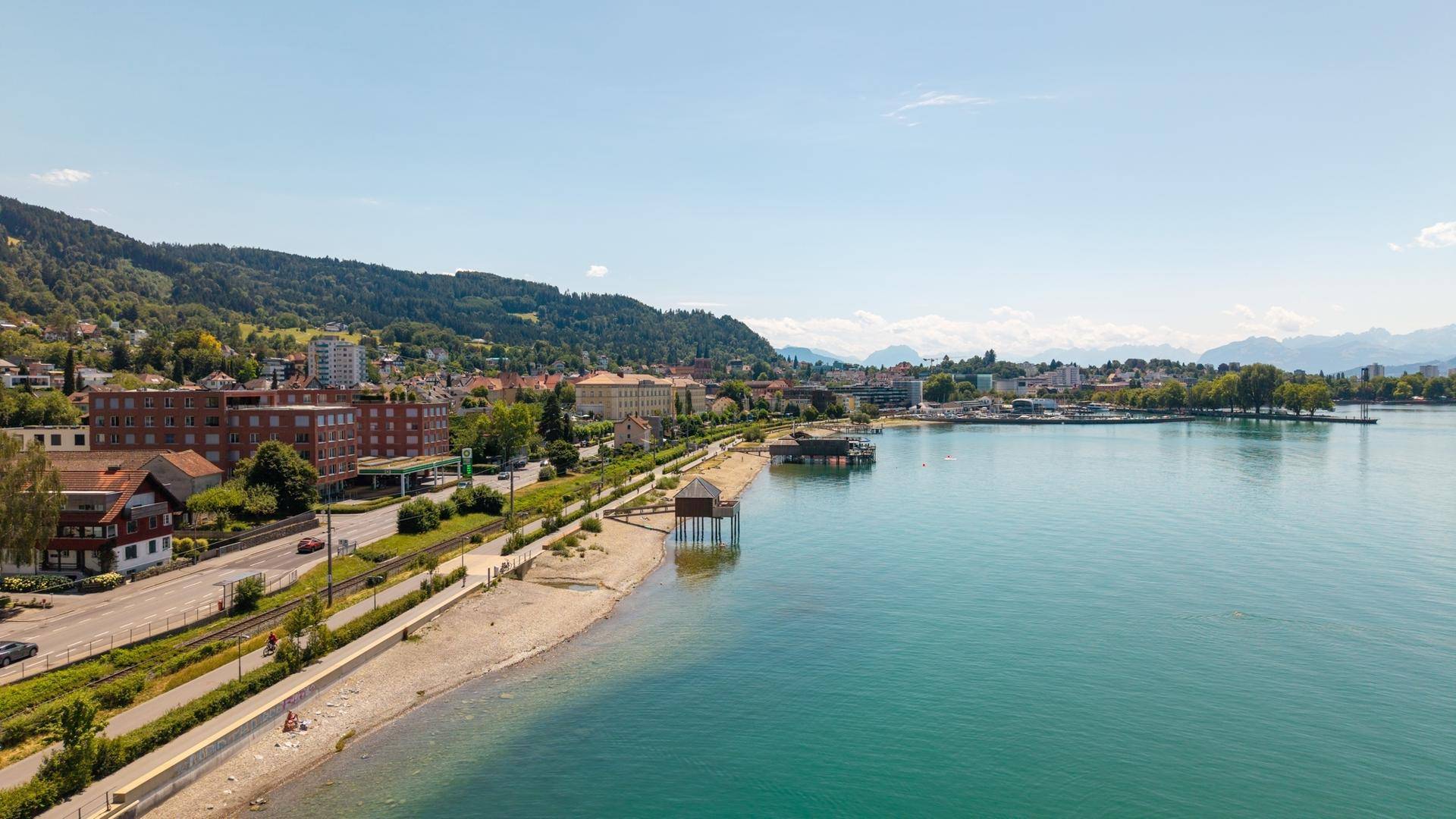 Luftaufnahme der Küstenstadt mit See, Strand und bewaldeten Hügeln unter strahlend blauem Himmel.