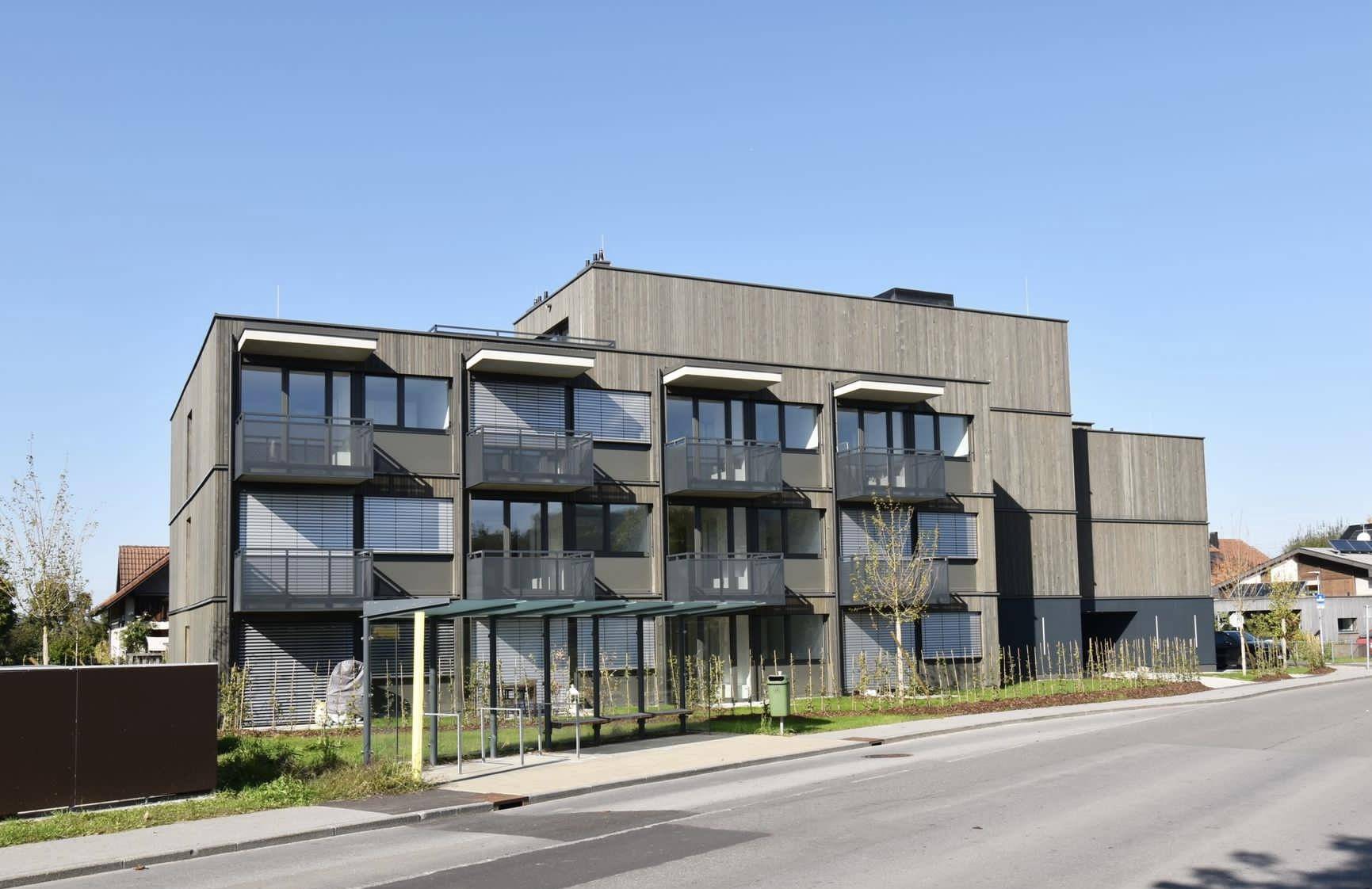 Modernes Mehrfamilienhaus mit Holzfassade, Balkonen und großen Fenstern in sonniger Umgebung.