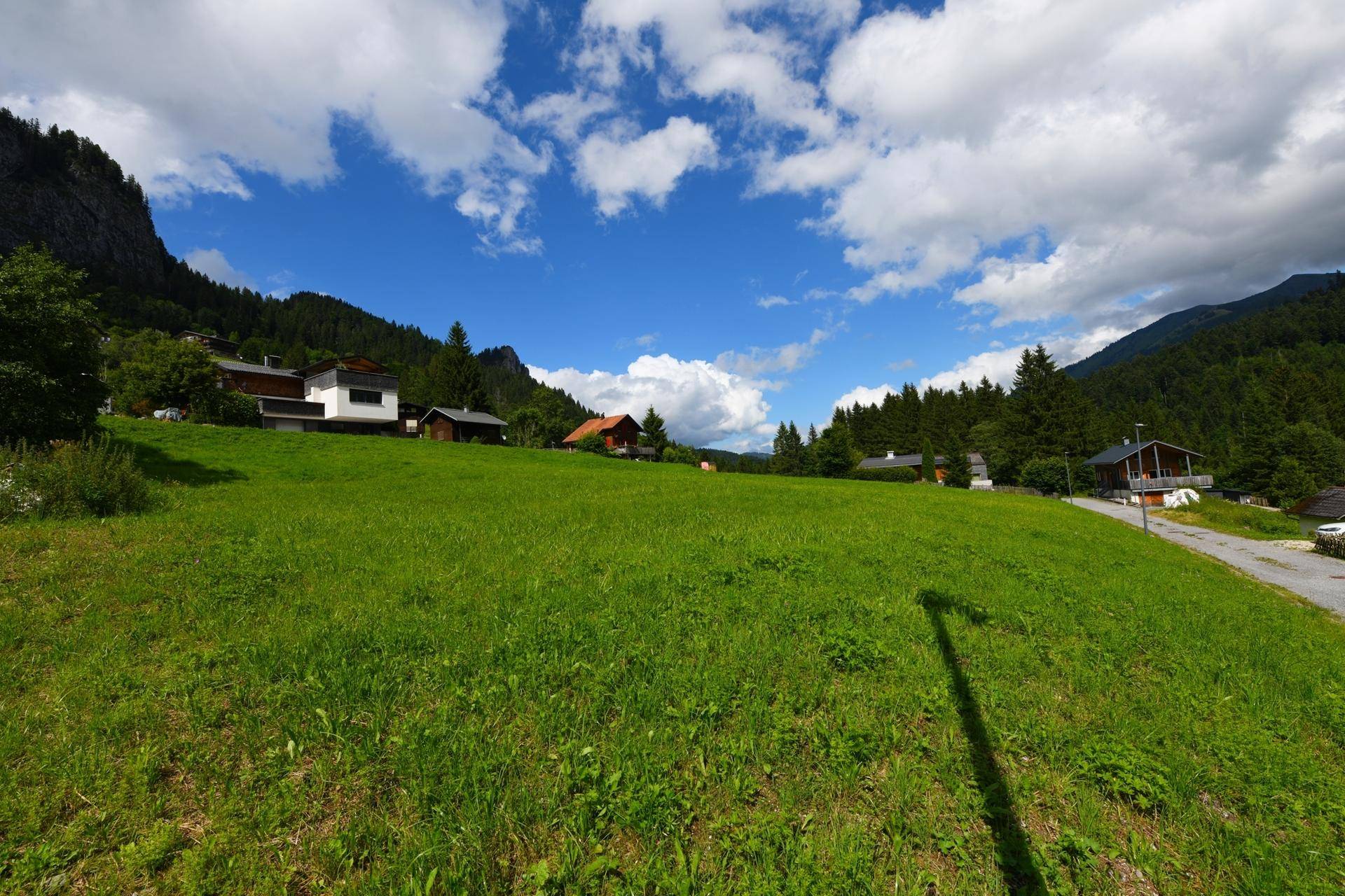 Panoramablick über eine grüne Wiese mit Häusern und majestätischen Bergen unter blauem Himmel.