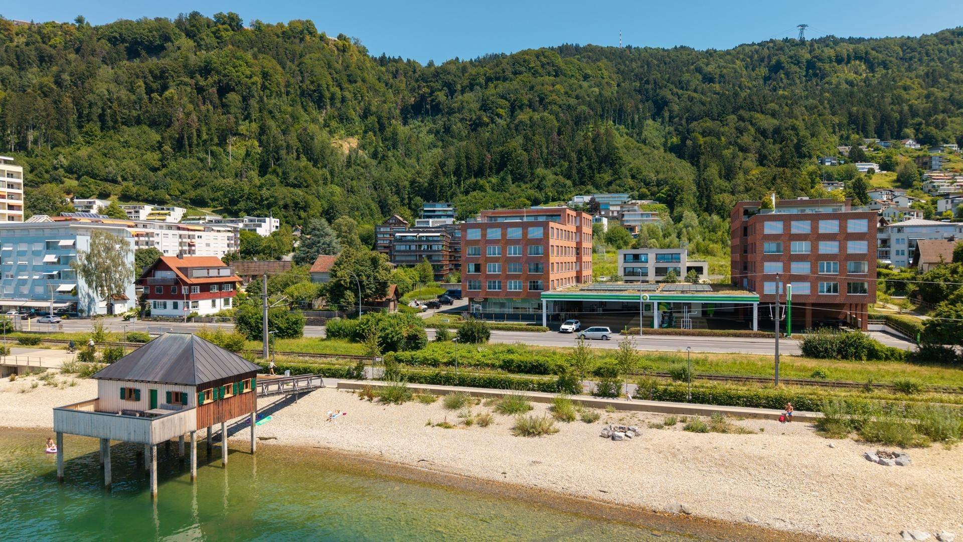 Blick auf den Strand mit einem Gebäude auf Stelzen, einer Tankstelle und bewaldeten Hügeln im Hintergrund.