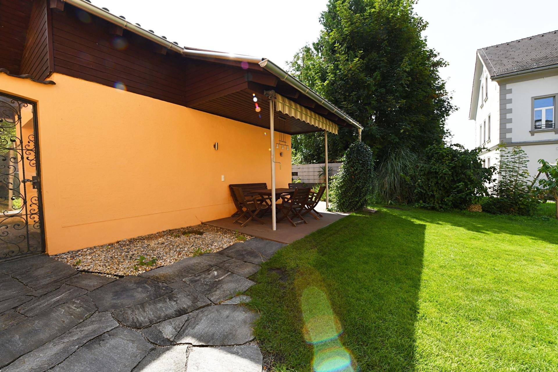 Garten mit Rasenfläche, Steinweg und überdachtem Sitzbereich im Freien.