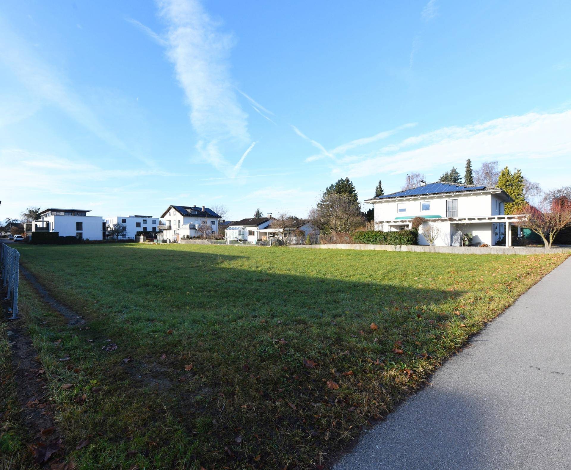 Weitläufiges grünes Grundstück unter blauem Himmel, umgeben von Wohnhäusern und einer Zufahrtsstraße.