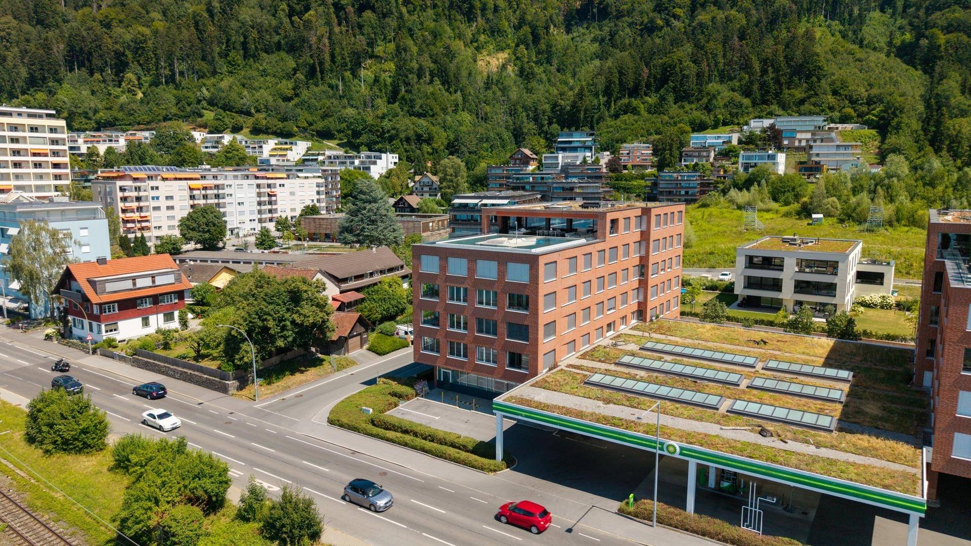 Luftaufnahme von modernen Wohngebäuden und einer Tankstelle mit begrüntem Dach am Fuße eines Hügels.
