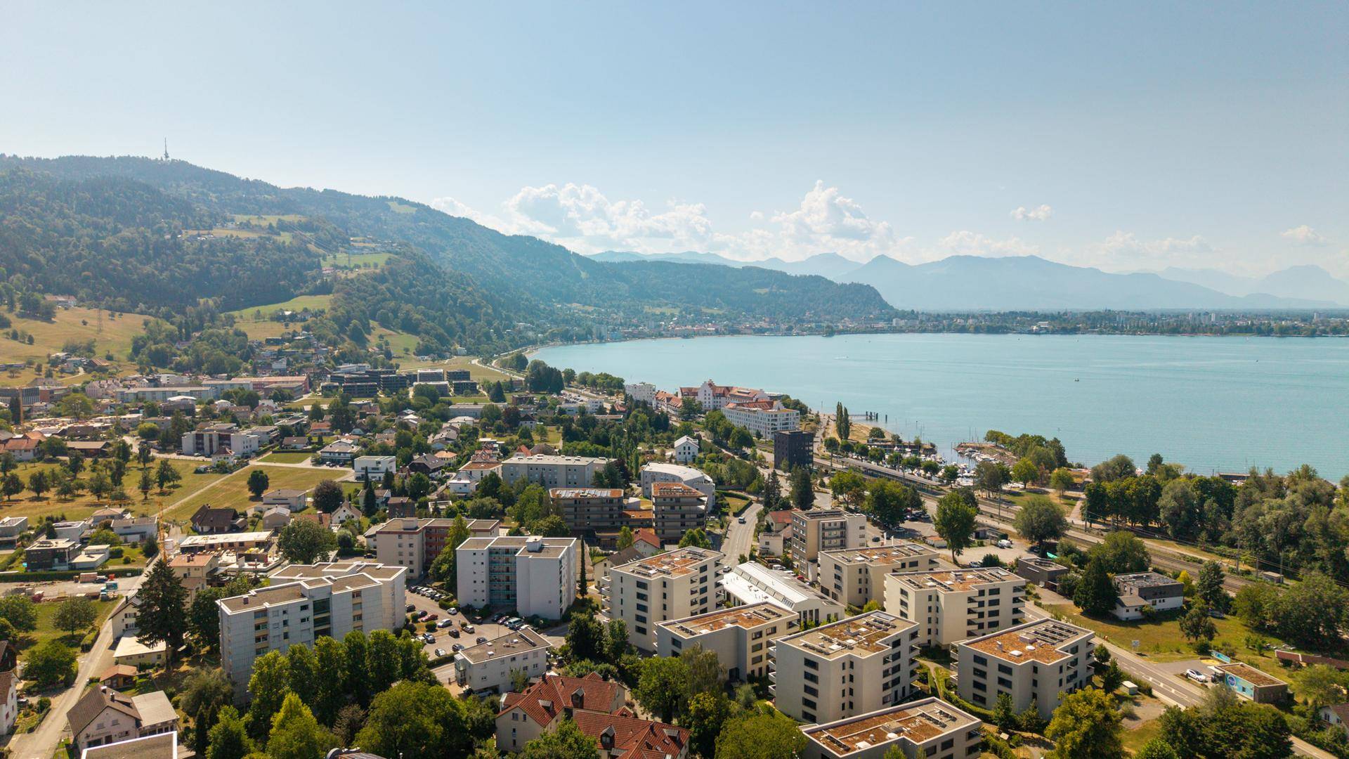 Panoramablick auf eine Stadt am Seeufer mit Bergen im Hintergrund und klarem blauen Wasser.