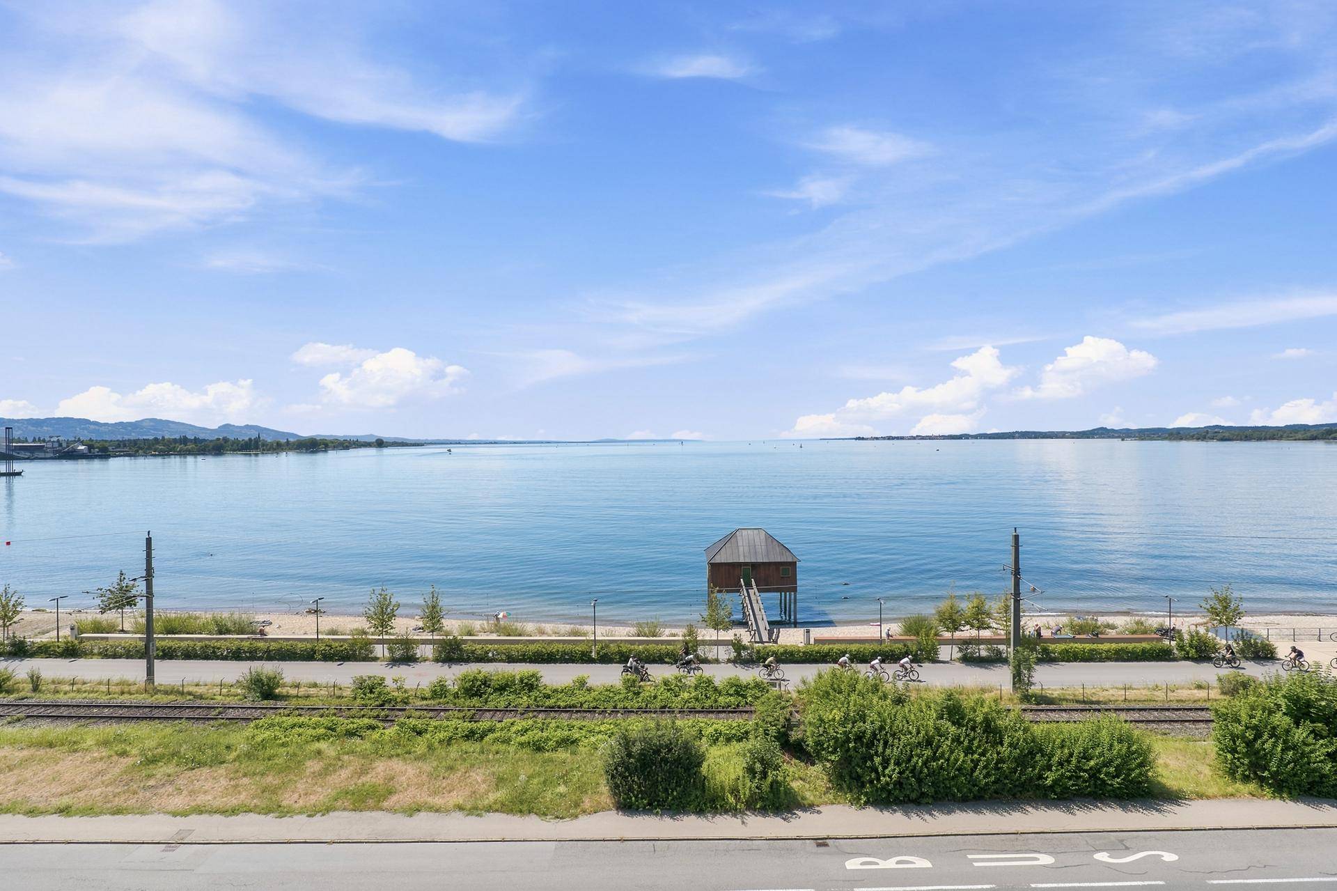 Weitläufiger Blick auf den See, den Strand und die umliegende Landschaft mit Bergen im Hintergrund.