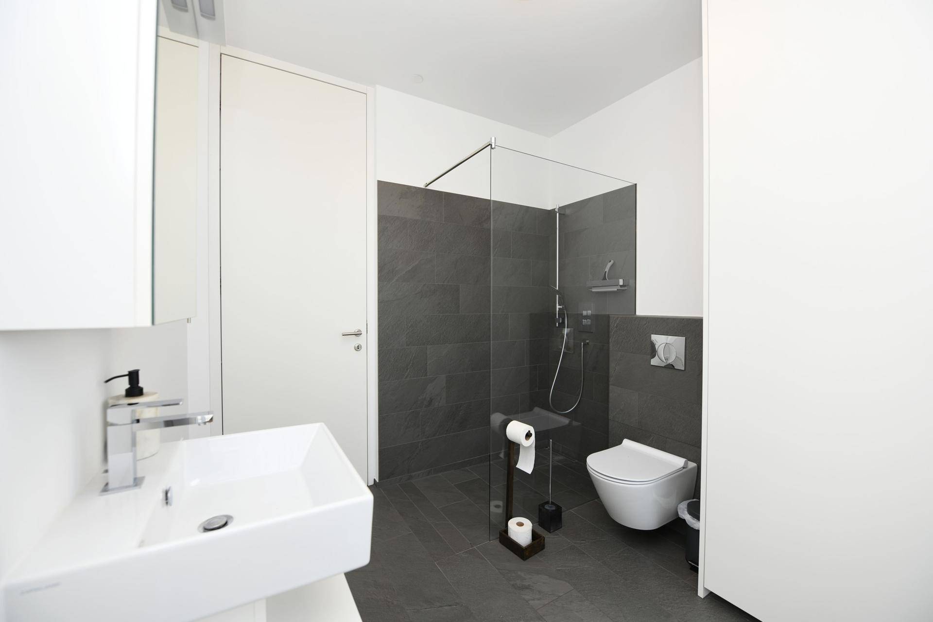 Helles Badezimmer mit modernem Waschbecken, ebenerdiger Dusche und dunklen Fliesen.