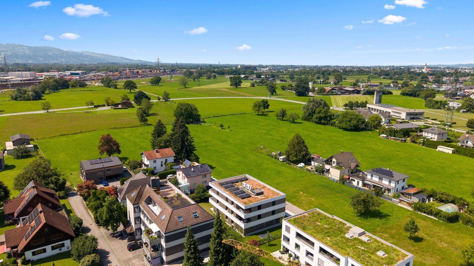 Panoramablick aus der Luft über eine weite grüne Landschaft mit Wohngebäuden und Bergen am Horizont.