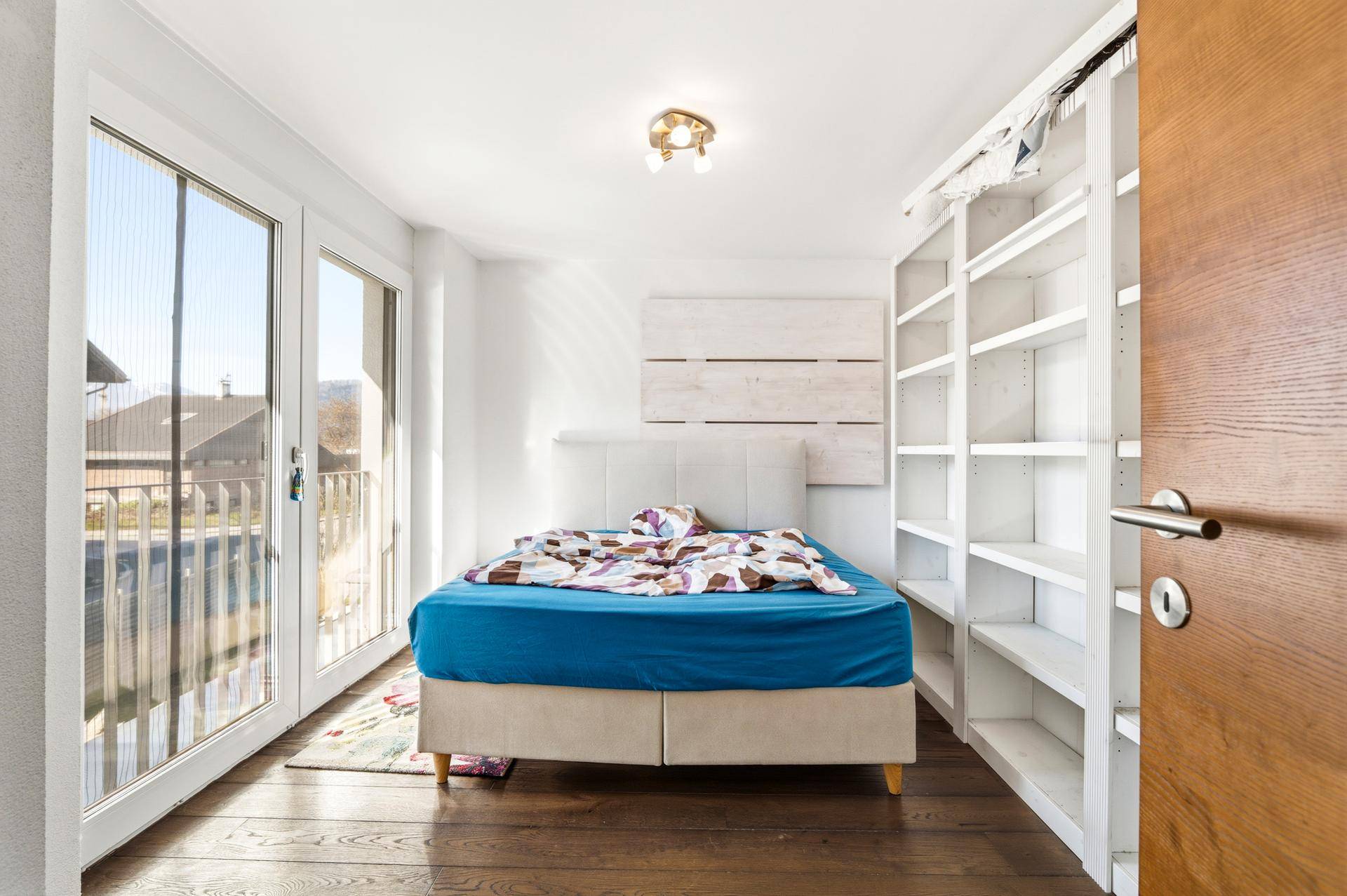 Helles Schlafzimmer mit Holzboden, Bett und Bücherregal, mit Balkonzugang.