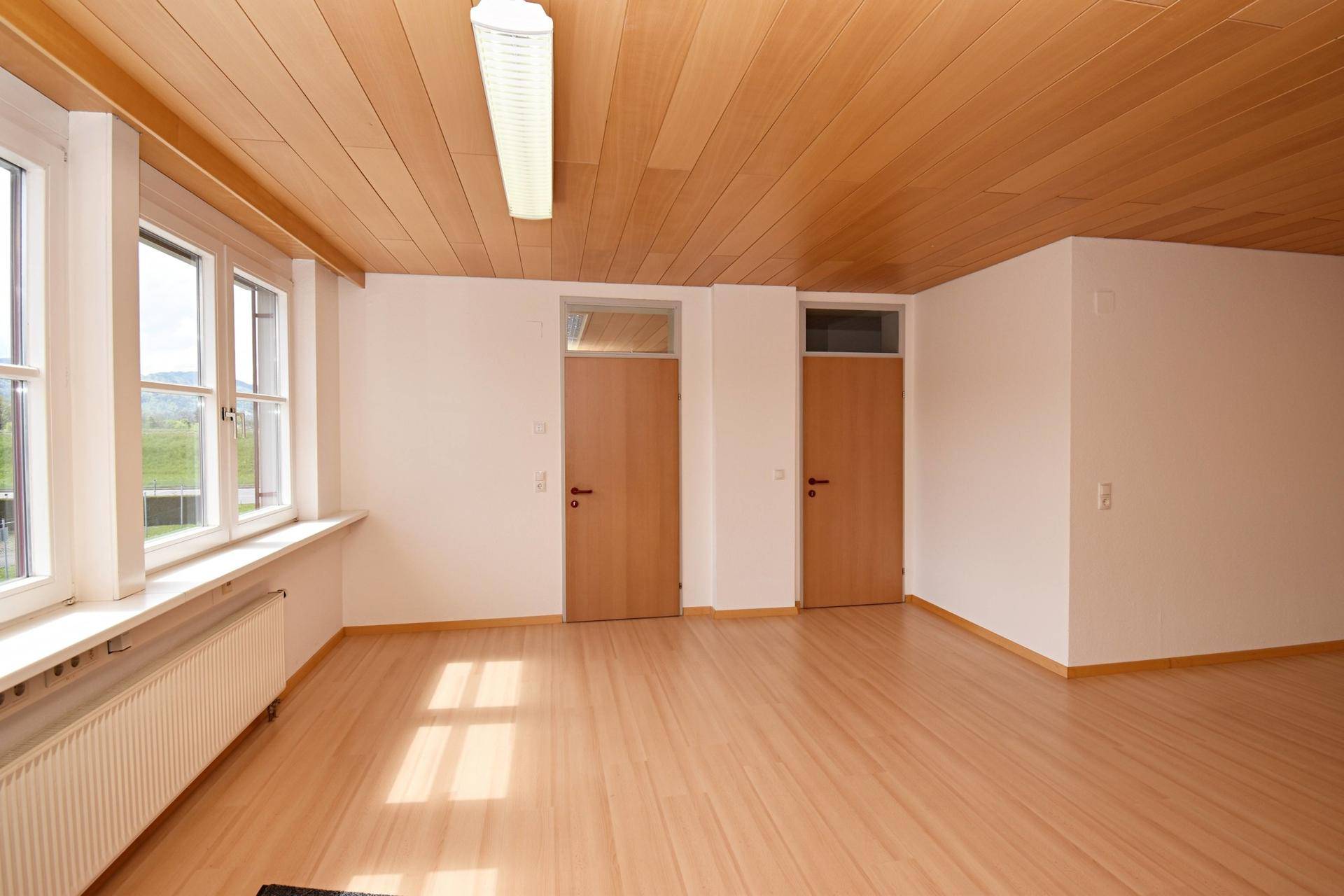 Helles, leeres Büro mit Holzdeckenverkleidung, Laminatboden und großen Fenstern.