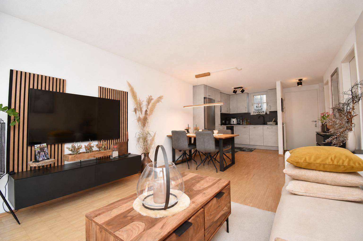 Moderner Wohnbereich mit TV-Wand, Essplatz und offener Küche, alles in einem hellen Ambiente.