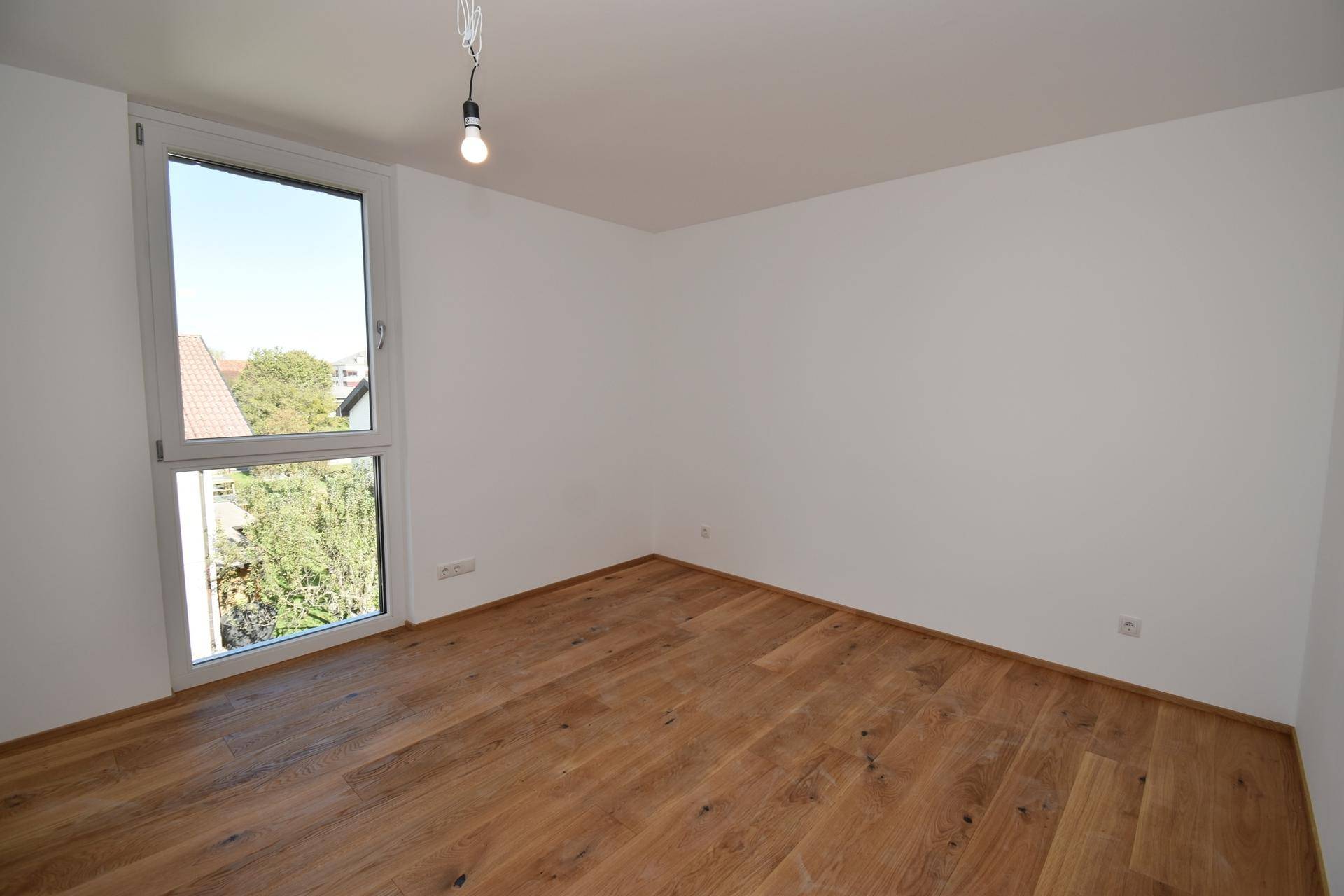 Schlafzimmer mit großem Fenster, das viel Tageslicht hereinlässt und Holzparkettboden.