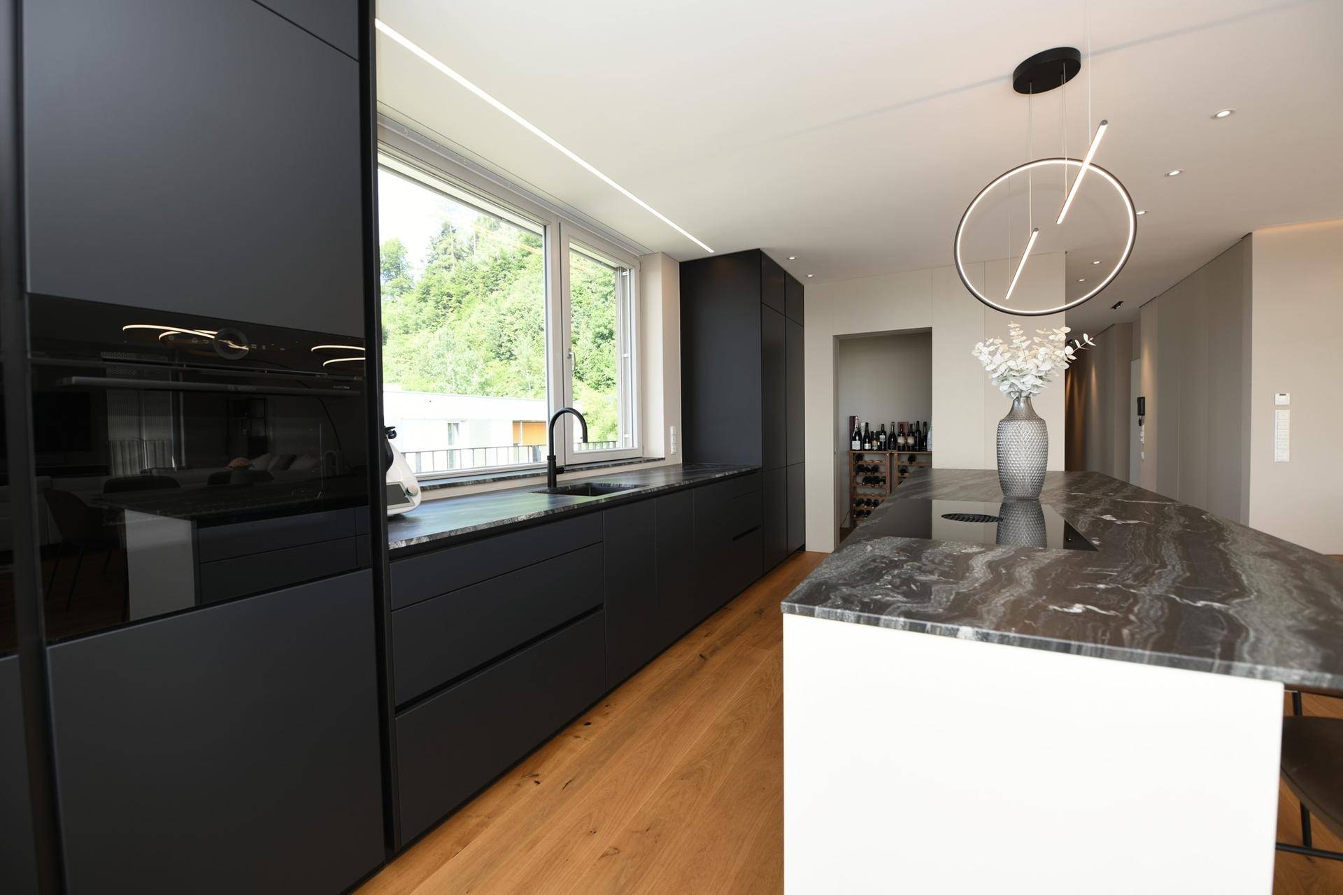 Moderne schwarze Küche mit integrierten Geräten, Fenster und Zugang zu einem Weinregal.