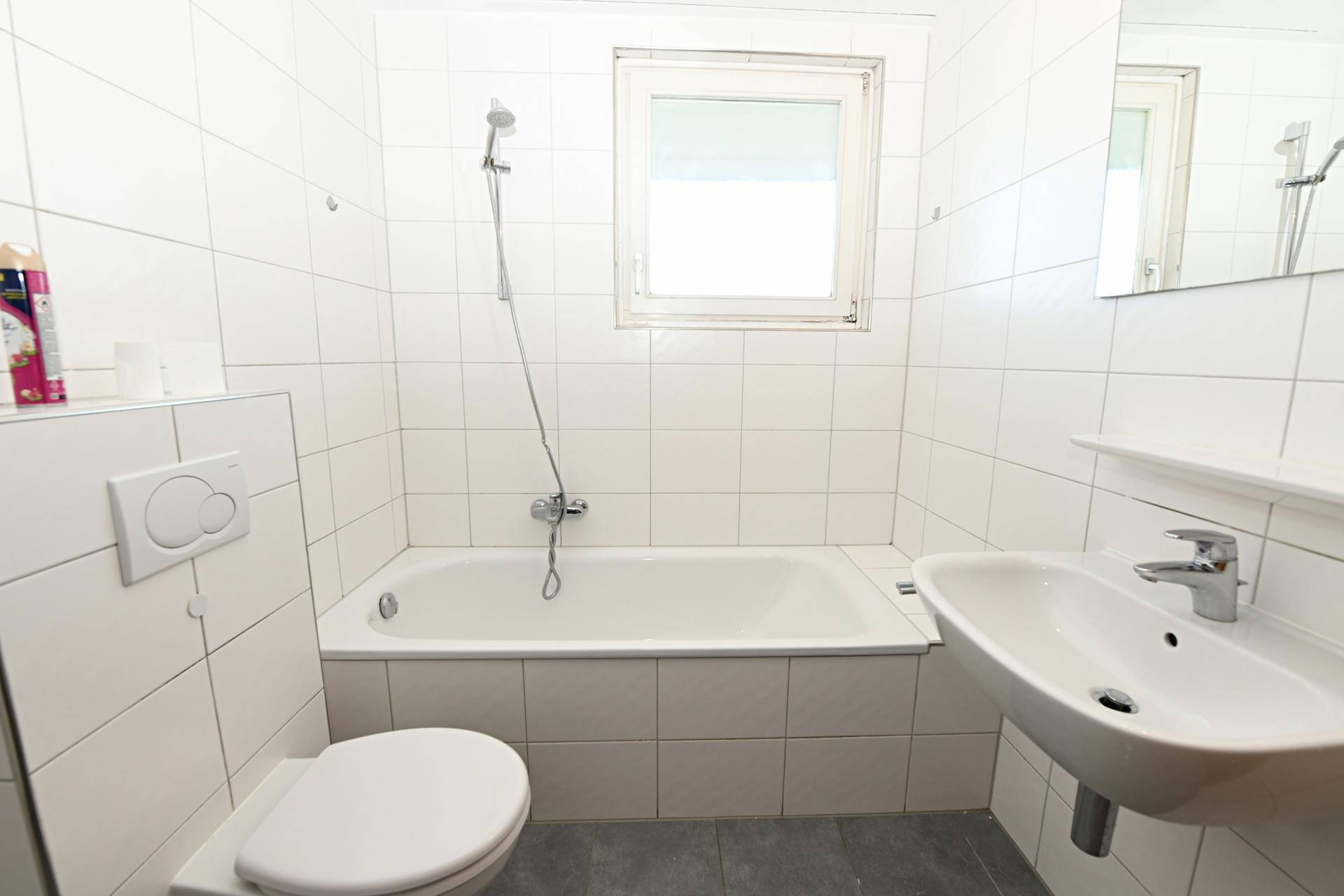 Funktionales Badezimmer mit weißen Fliesen, Badewanne, WC, Waschbecken und Fenster.