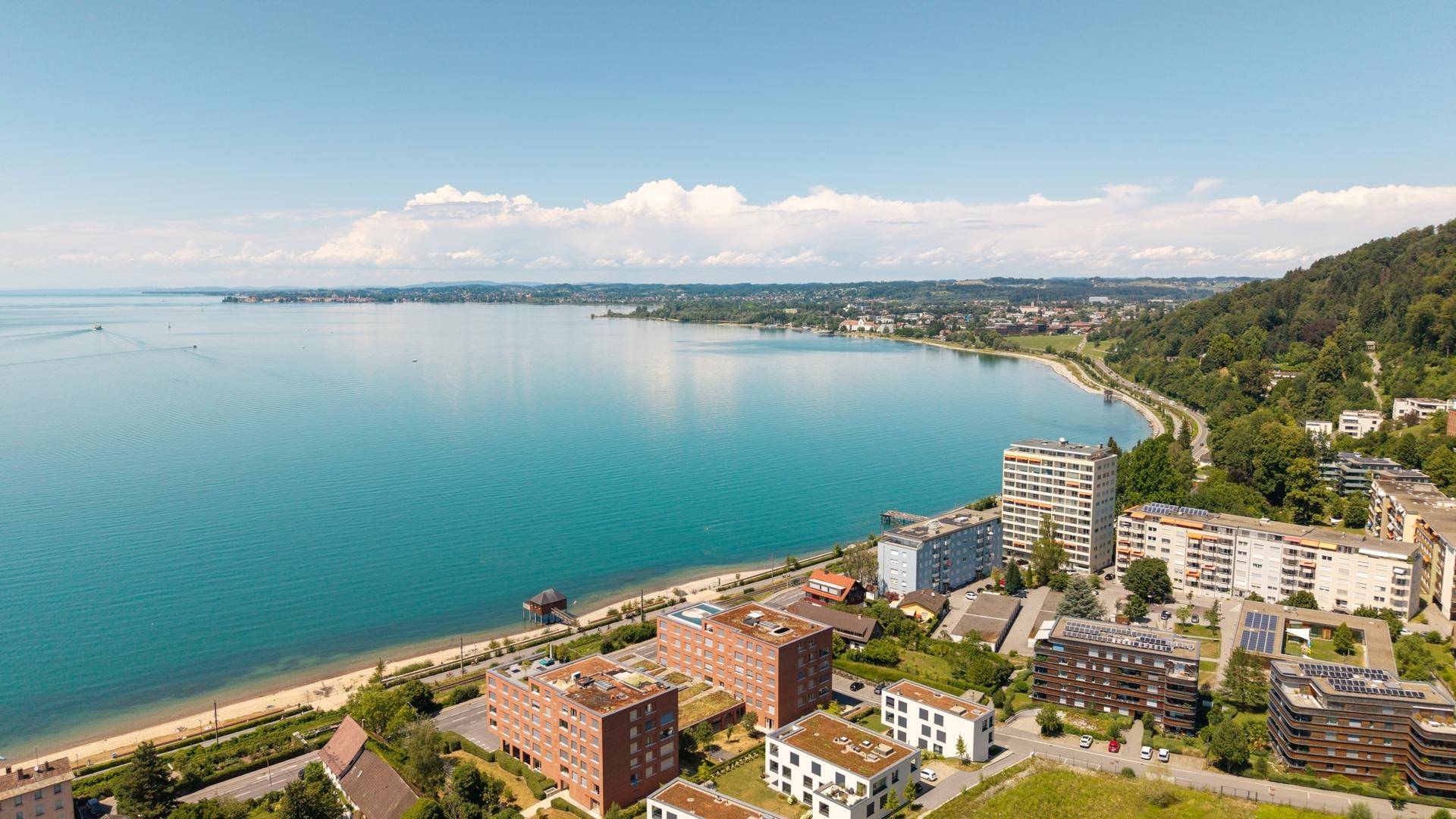 Panoramablick auf die Stadt am Seeufer mit umliegenden Bergen und Wohngebäuden unter blauem Himmel.