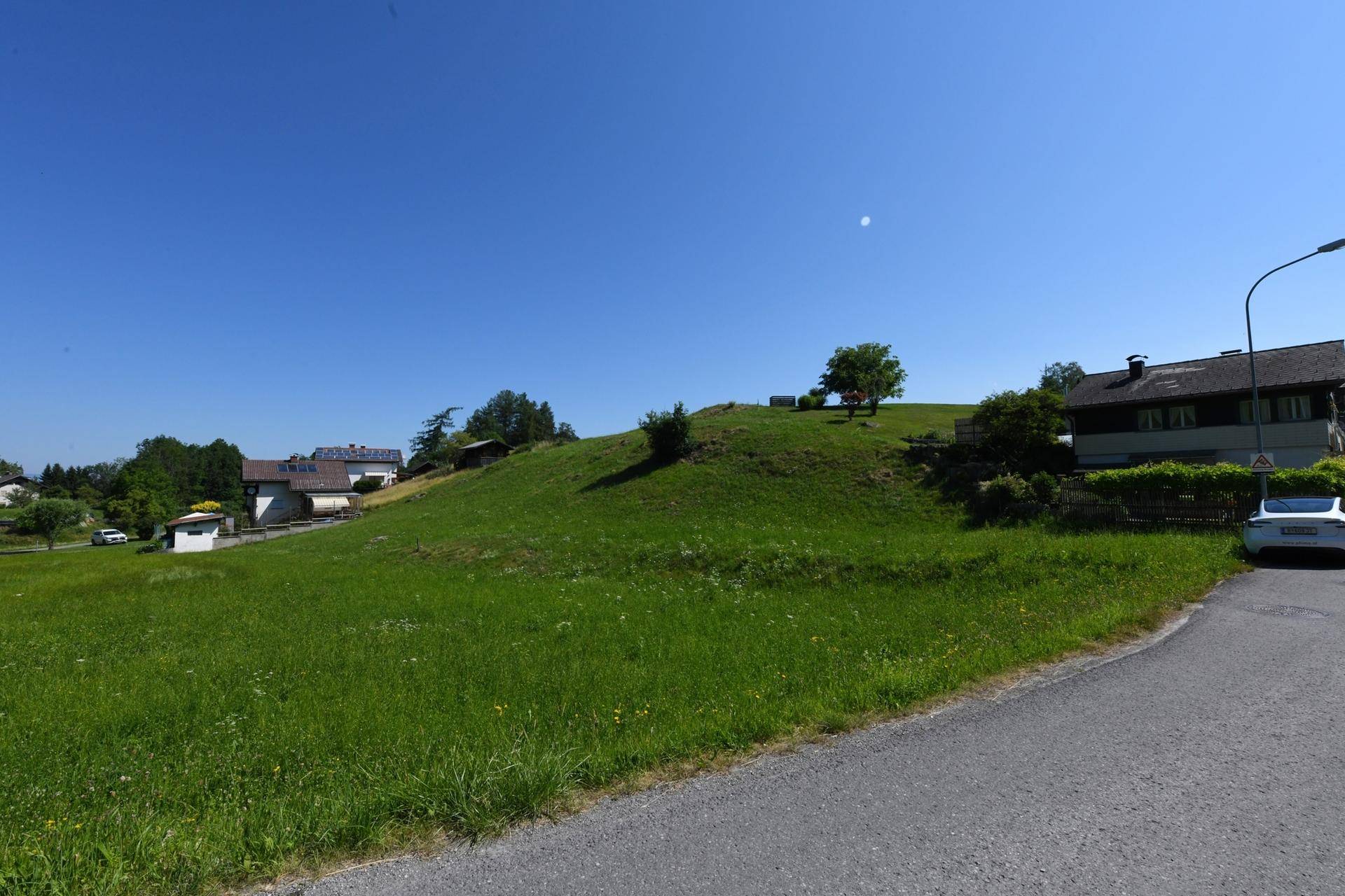 Sonniger Blick auf ein grünes Hanggrundstück mit Straße im Vordergrund und Häusern im Hintergrund.