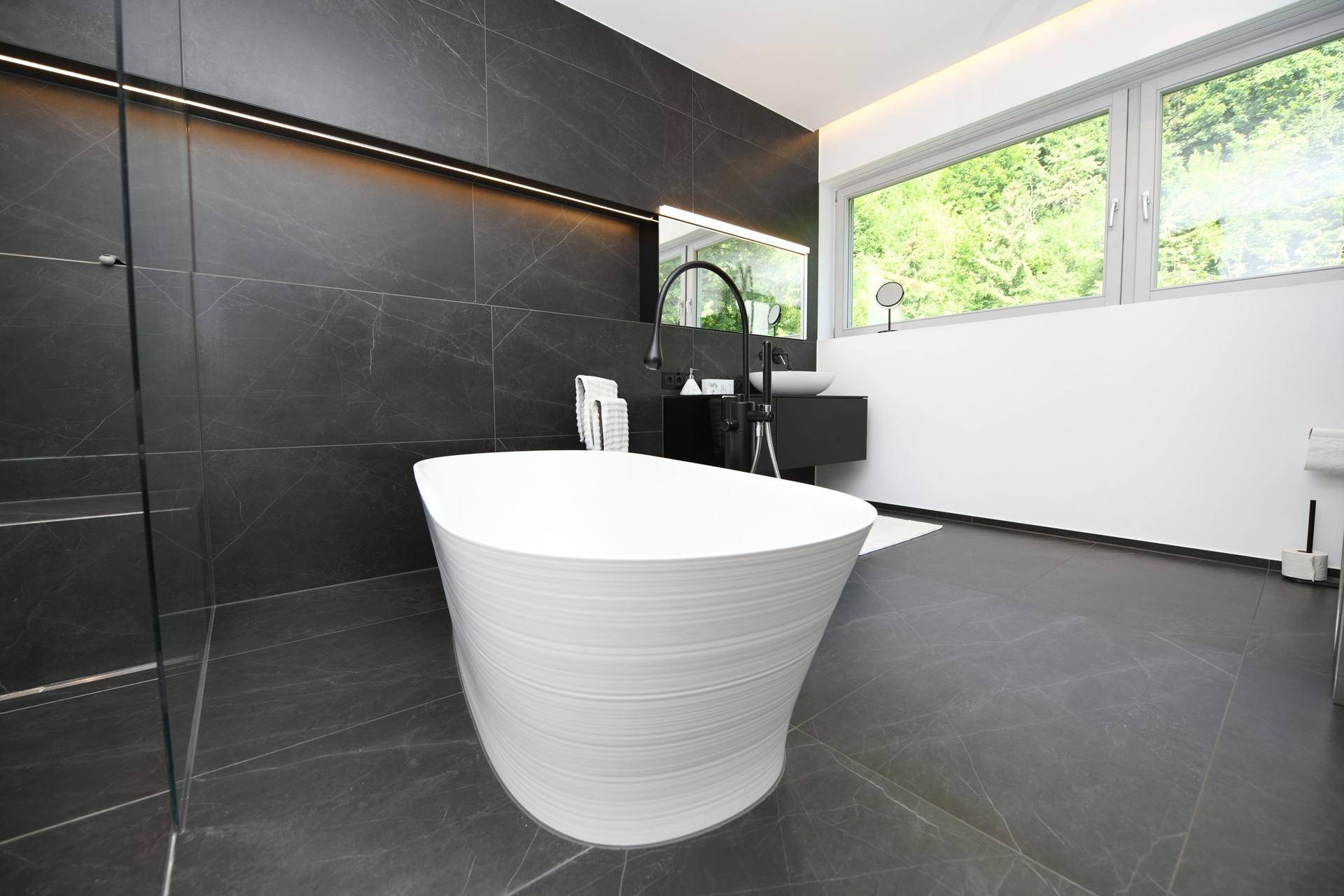 Elegantes Badezimmer mit freistehender Badewanne, dunklen Fliesen und großem Fenster.