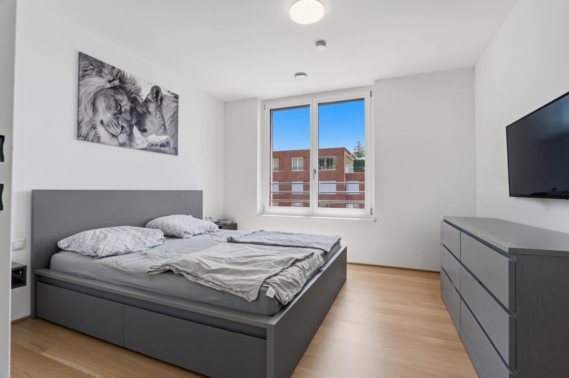 Modernes Schlafzimmer mit grauem Bett, Holzboden und großem Fenster für viel Tageslicht.
