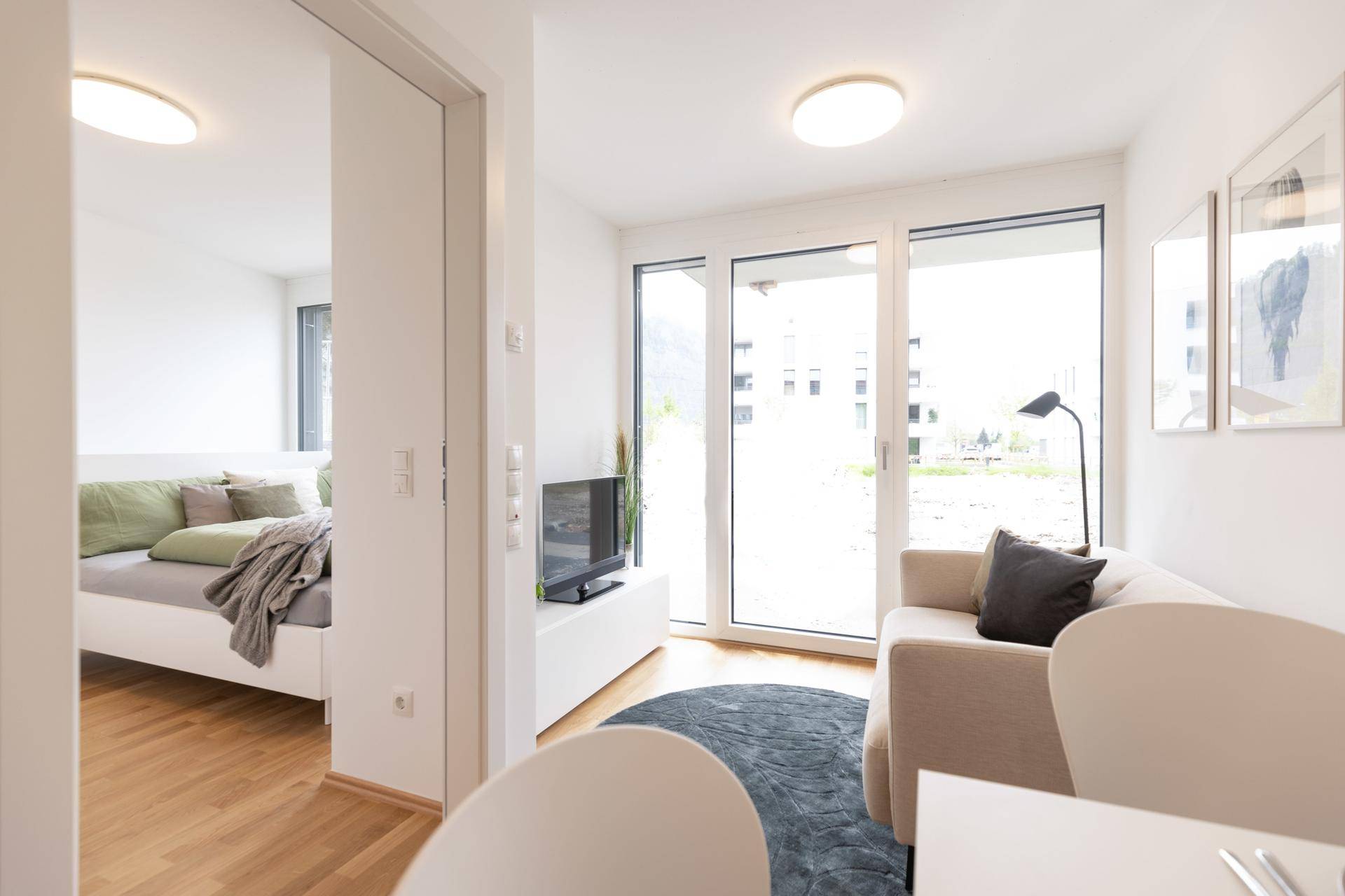 Heller Wohnbereich mit großen Fenstern und Blick in das angrenzende Schlafzimmer, modern eingerichtet.