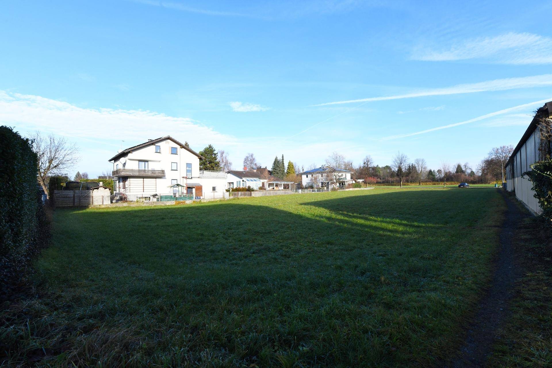 Sonniges, offenes Grundstück mit Rasenfläche und Wohnhäusern im Hintergrund unter klarem Himmel.