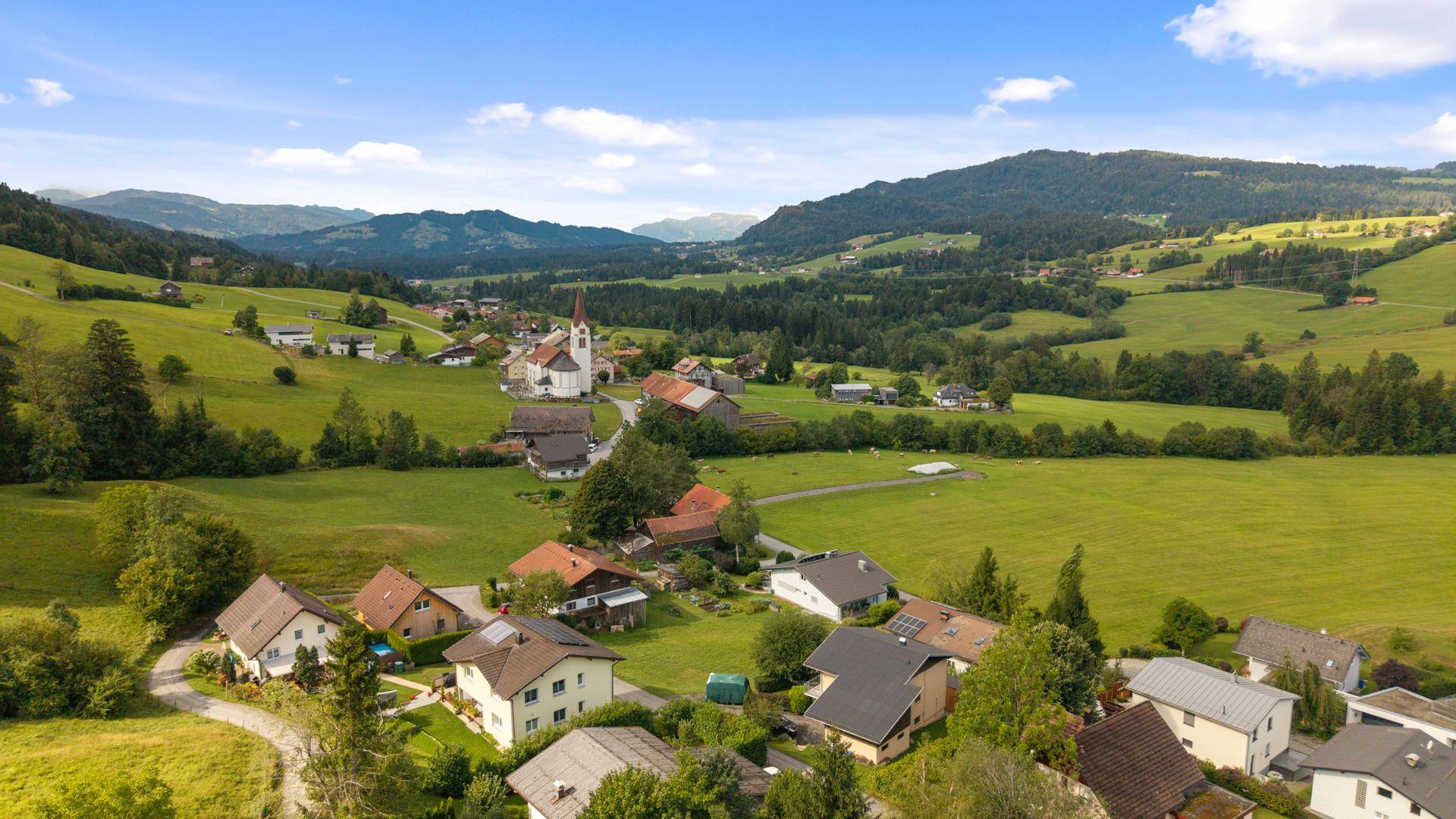 Malerische Luftaufnahme eines Dorfes mit Kirche, umgeben von weiten Feldern und sanften Hügeln.