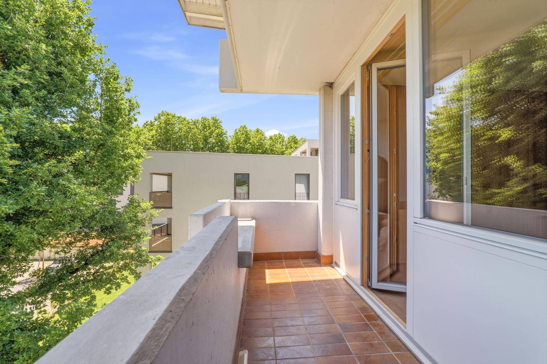 Balkon mit Fliesenboden und Blick auf Bäume und ein modernes Gebäude.