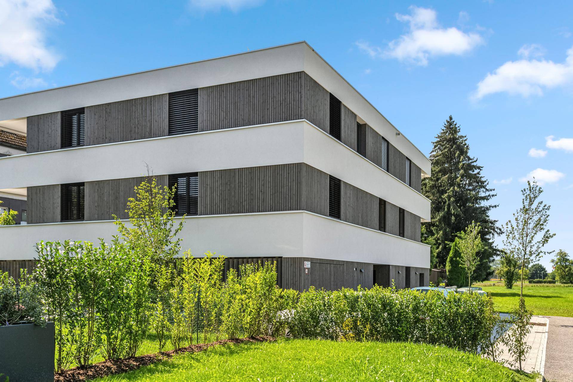 Moderne Gebäudefassade mit Holzverkleidung und großen Fenstern, umgeben von gepflegten Hecken.