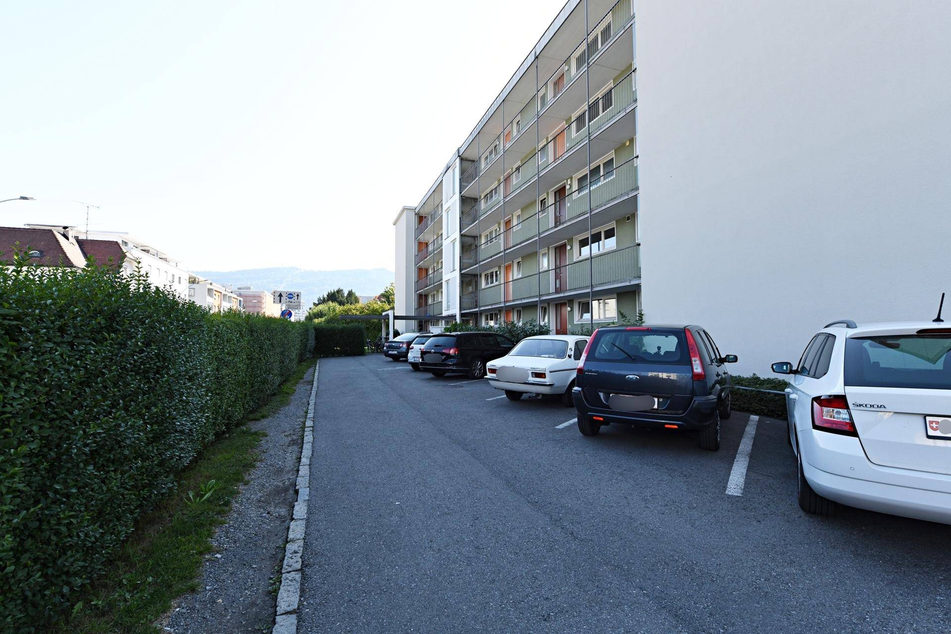 Mehrstöckiges Wohngebäude mit Außenparkplätzen und einer gepflegten Hecke entlang der Straße.