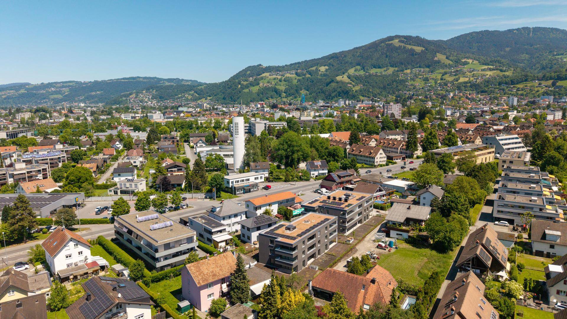 Weitläufige Luftaufnahme einer Stadt mit Wohngebäuden, grünen Hügeln und Bergen im Hintergrund unter blauem Himmel.